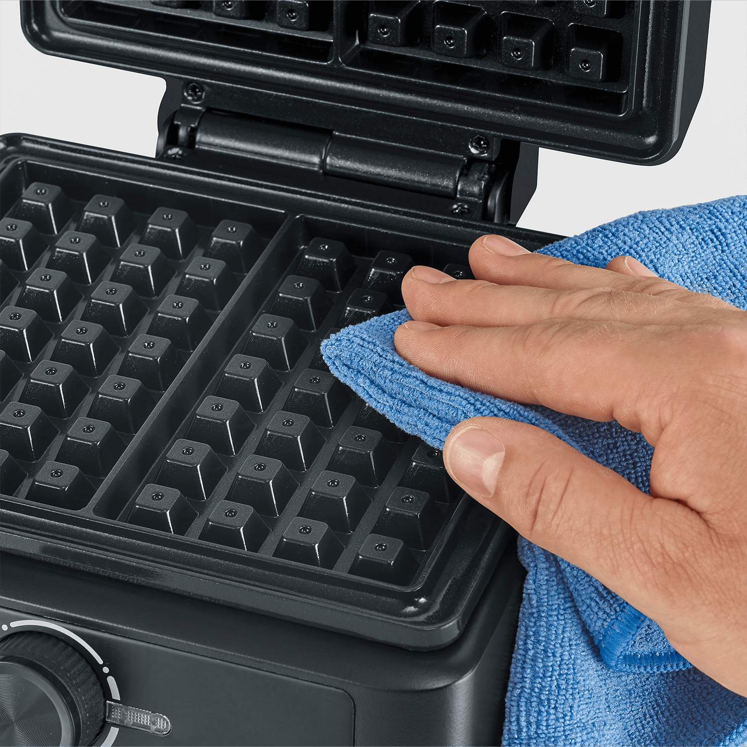 Eine Hand reinigt ein offenes schwarzes Waffeleisen mit einem blauen Tuch.