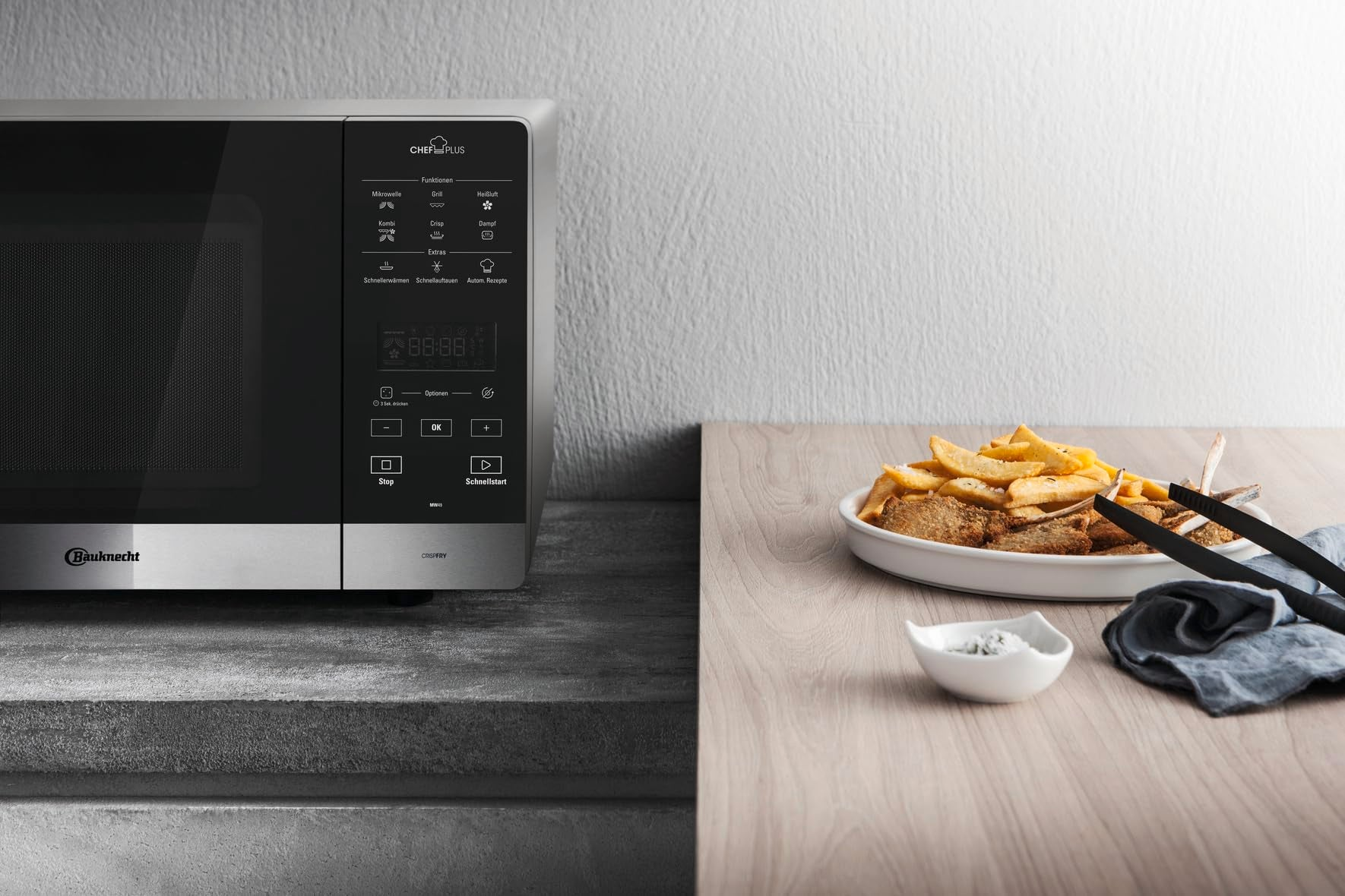 Micro-ondes moderne à gauche; assiette de frites et de viande avec des pinces sur table en bois à droite.