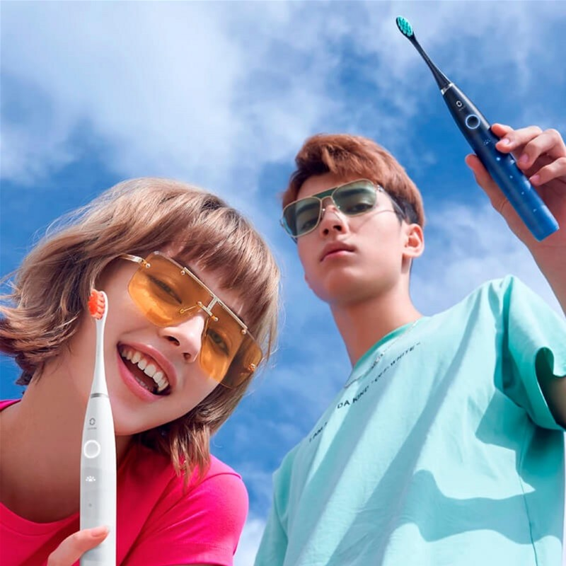 Ein junges Paar mit Sonnenbrille hält elektrische Zahnbürsten vor blauem Himmel.