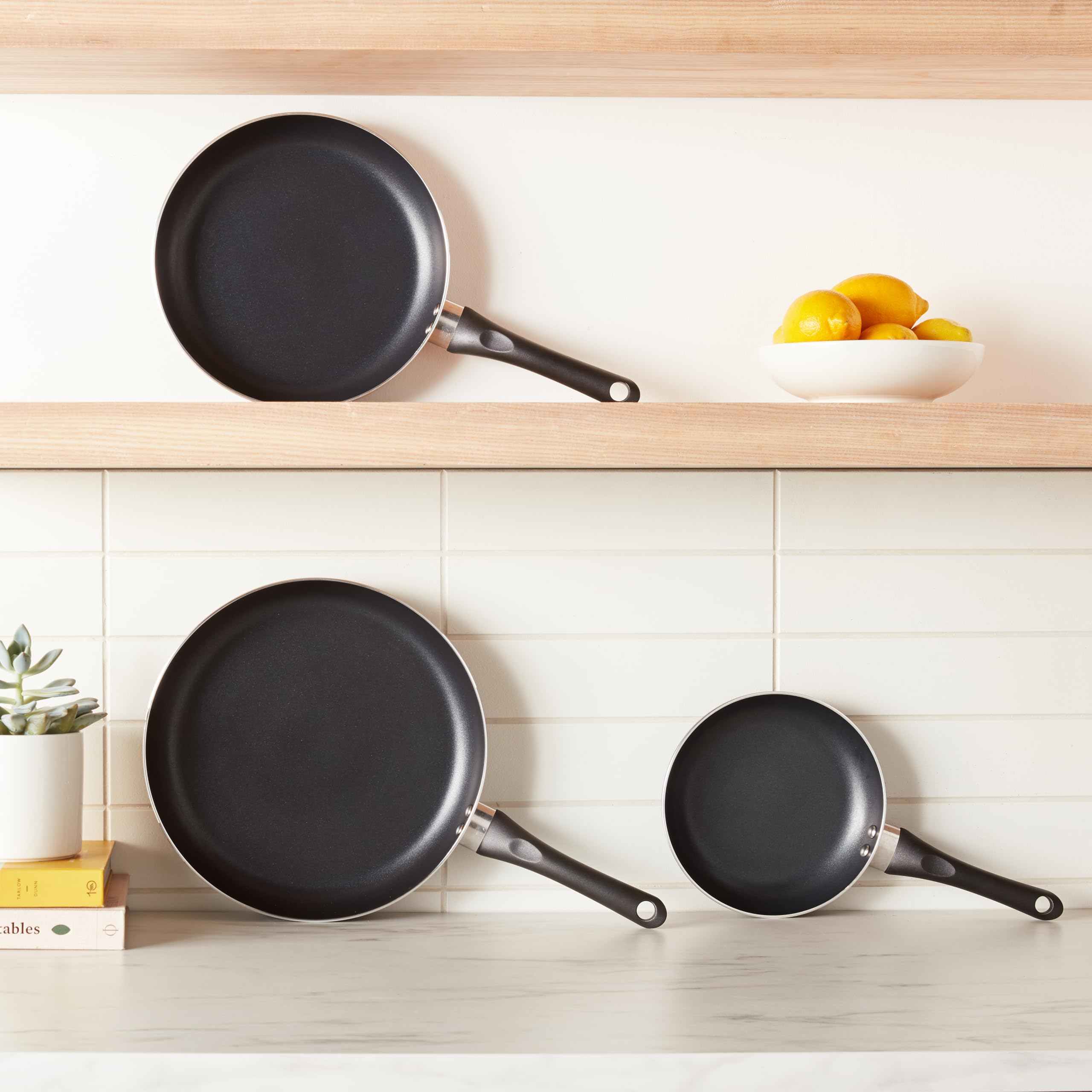 Trois poêles antiadhésives noires sur un comptoir et étagère, avec des citrons dans un bol et une plante en pot.