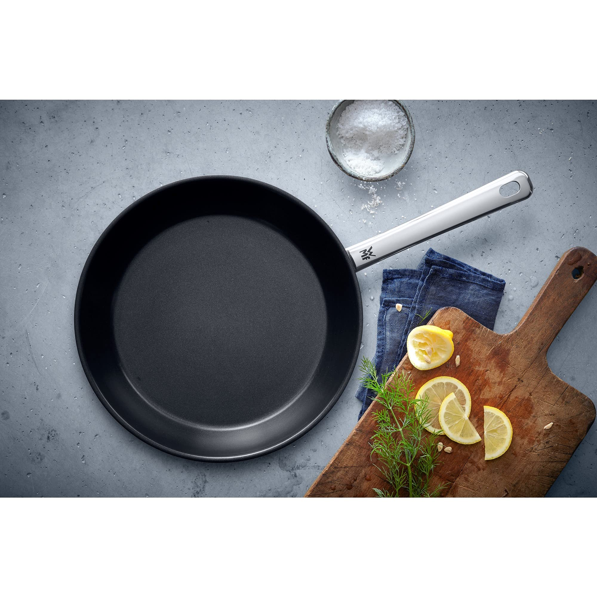 Une poêle noire avec manche argenté sur une surface grise, près d'une planche en bois avec citrons et herbes.