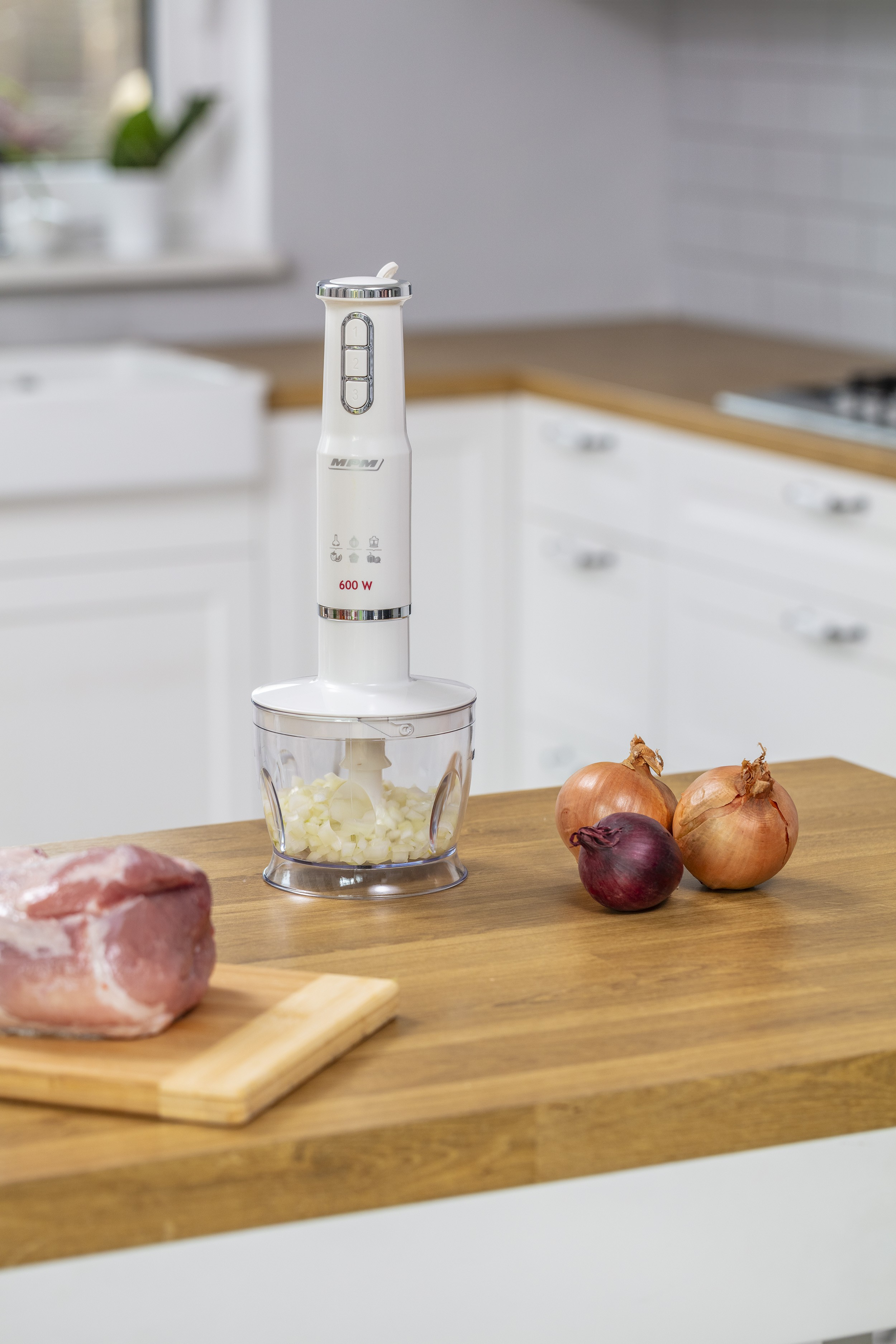 Biały blender ręczny z posiekaną cebulą w pojemniku stoi na drewnianym blacie kuchennym.
