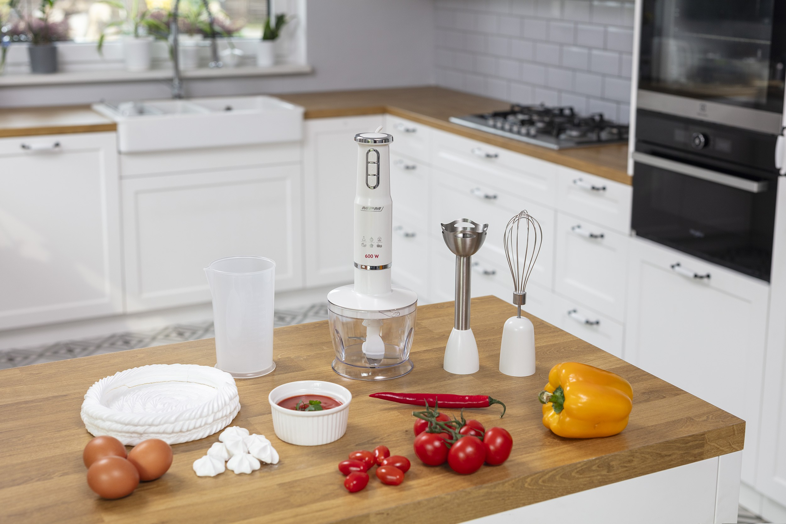Biały blender ręczny z akcesoriami oraz świeżą żywnością, taką jak pomidory, papryka i jajka, na wyspie kuchennej.