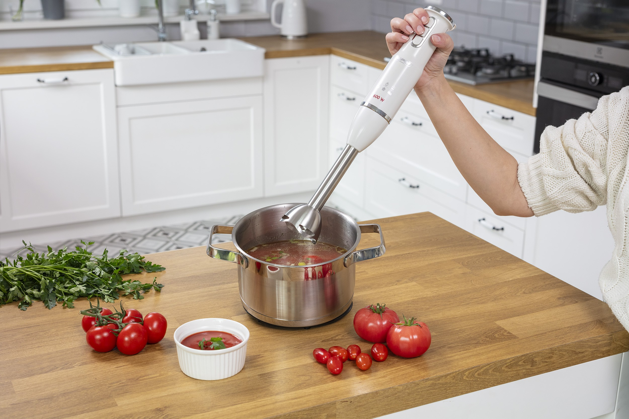 Ręka trzyma blender ręczny nad garnkiem z zupą na drewnianym blacie z pomidorami.