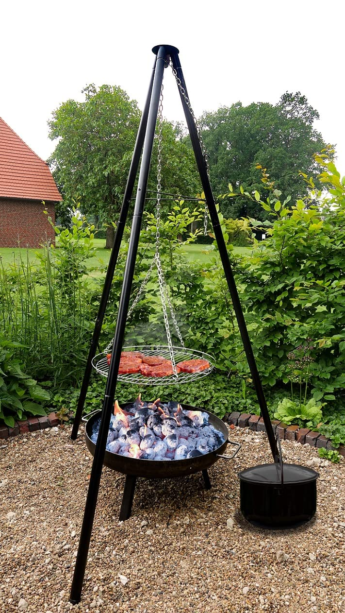 Ein dreibeiniger Grill mit hängendem Rost über glühenden Kohlen im Garten vor grünem Laub.