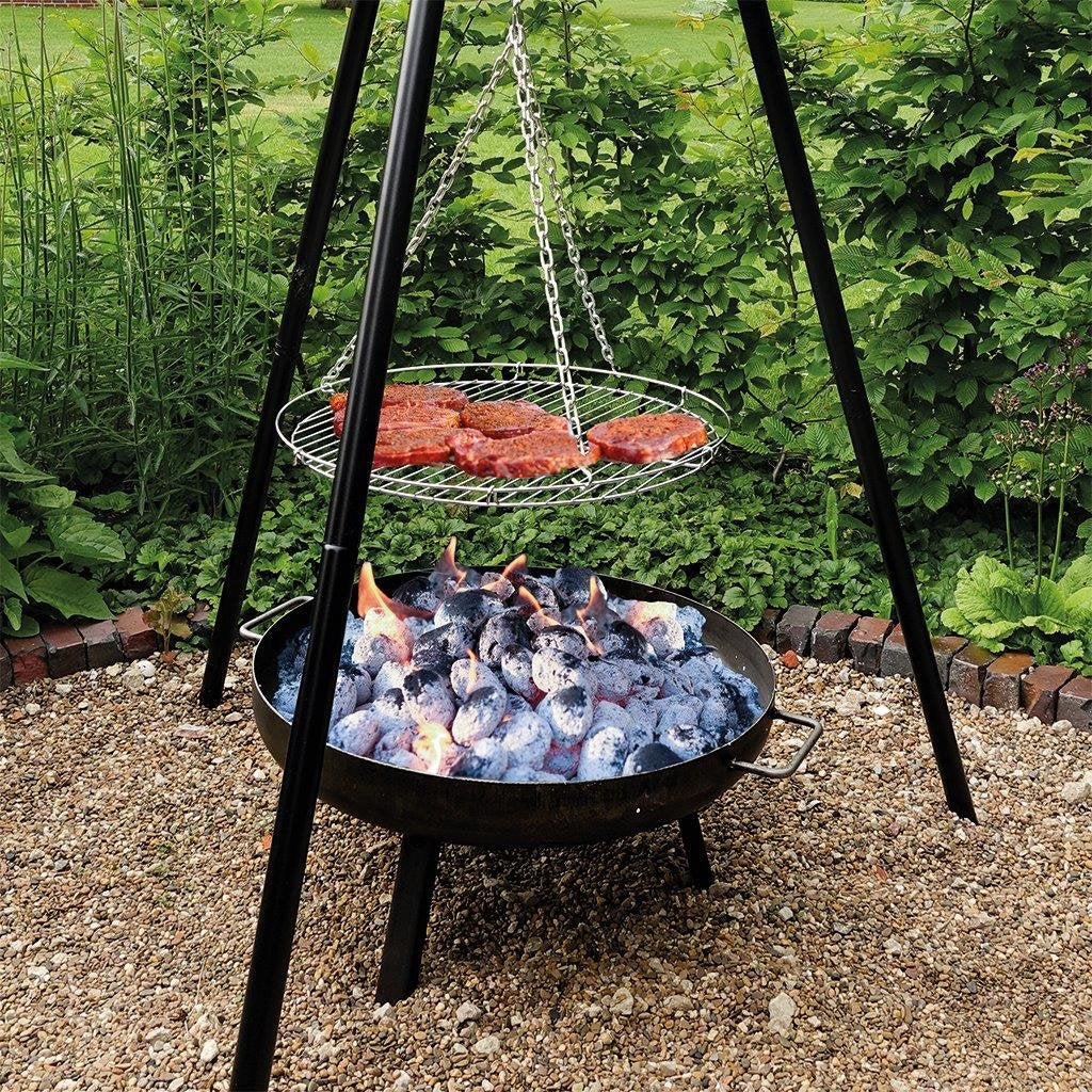 Ein Dreibein-Grill mit hängendem Rost und Steaks über einer Feuerschale im Garten.