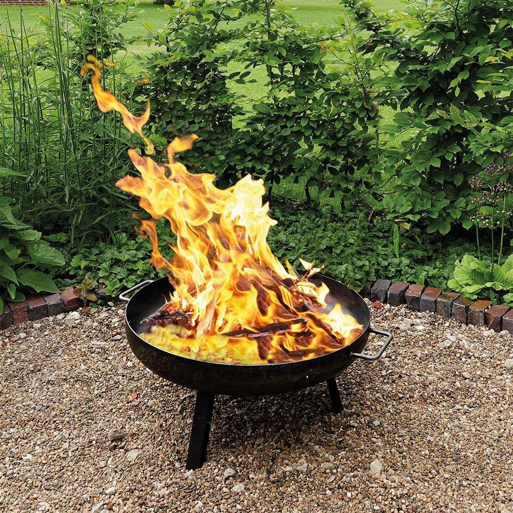 Eine schwarze Feuerschale auf Kies mit hellem, knisterndem Holzfeuer, vor dem Hintergrund grüner Büsche.