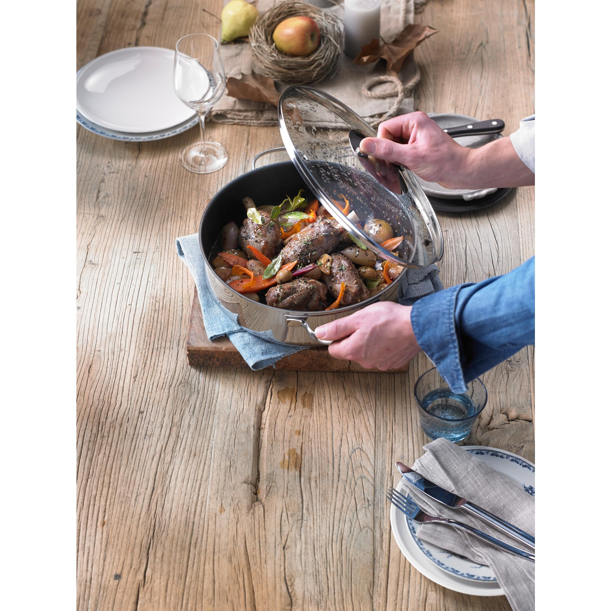 Eine Person hebt den Glasdeckel eines Topfes mit Fleisch, Karotten und Kartoffeln auf einem Holztisch an.