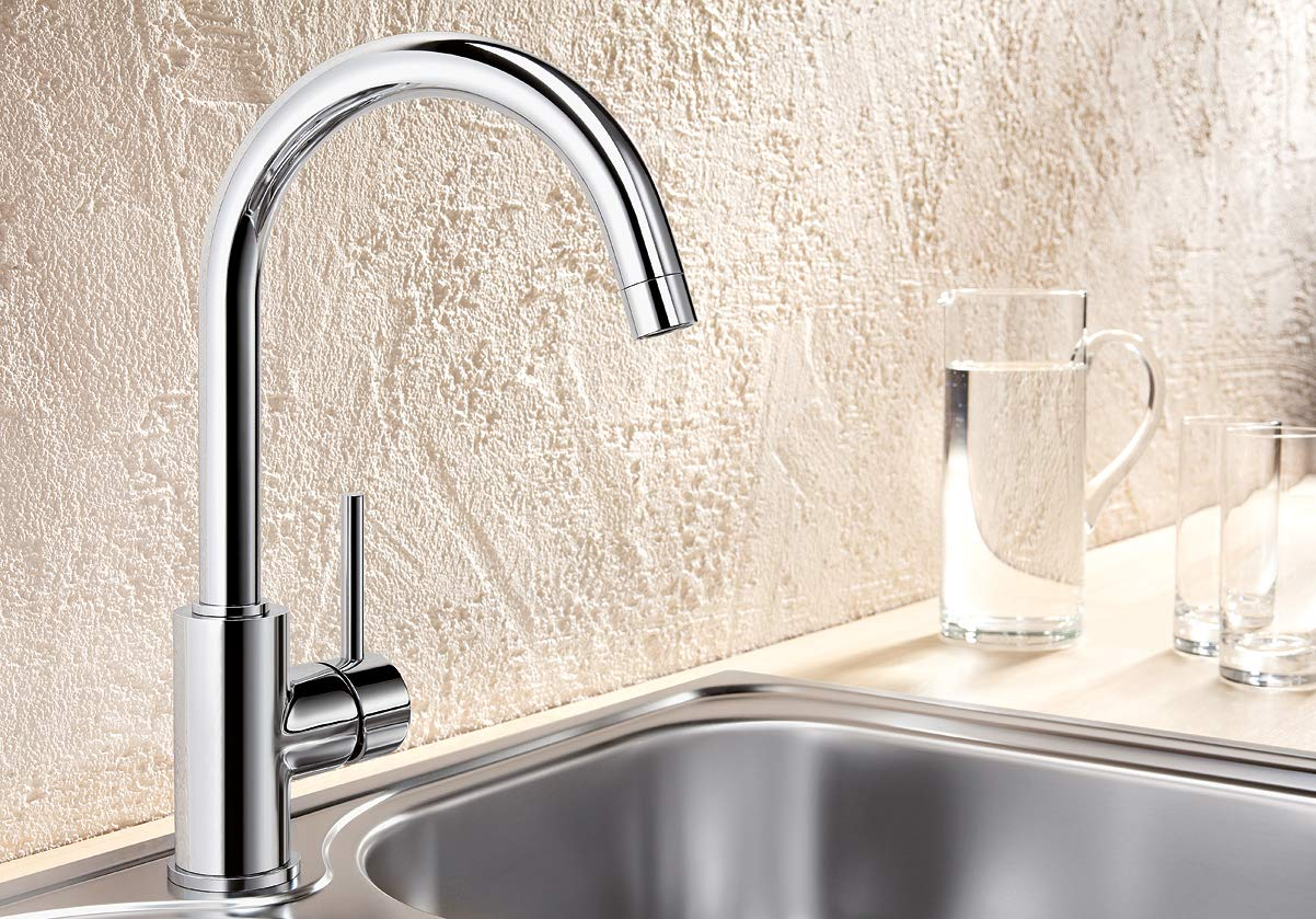 Glänzender Chrom-Wasserhahn an einer Edelstahlspüle vor einer strukturierten Wand, mit einer Wasserkanne im Hintergrund.
