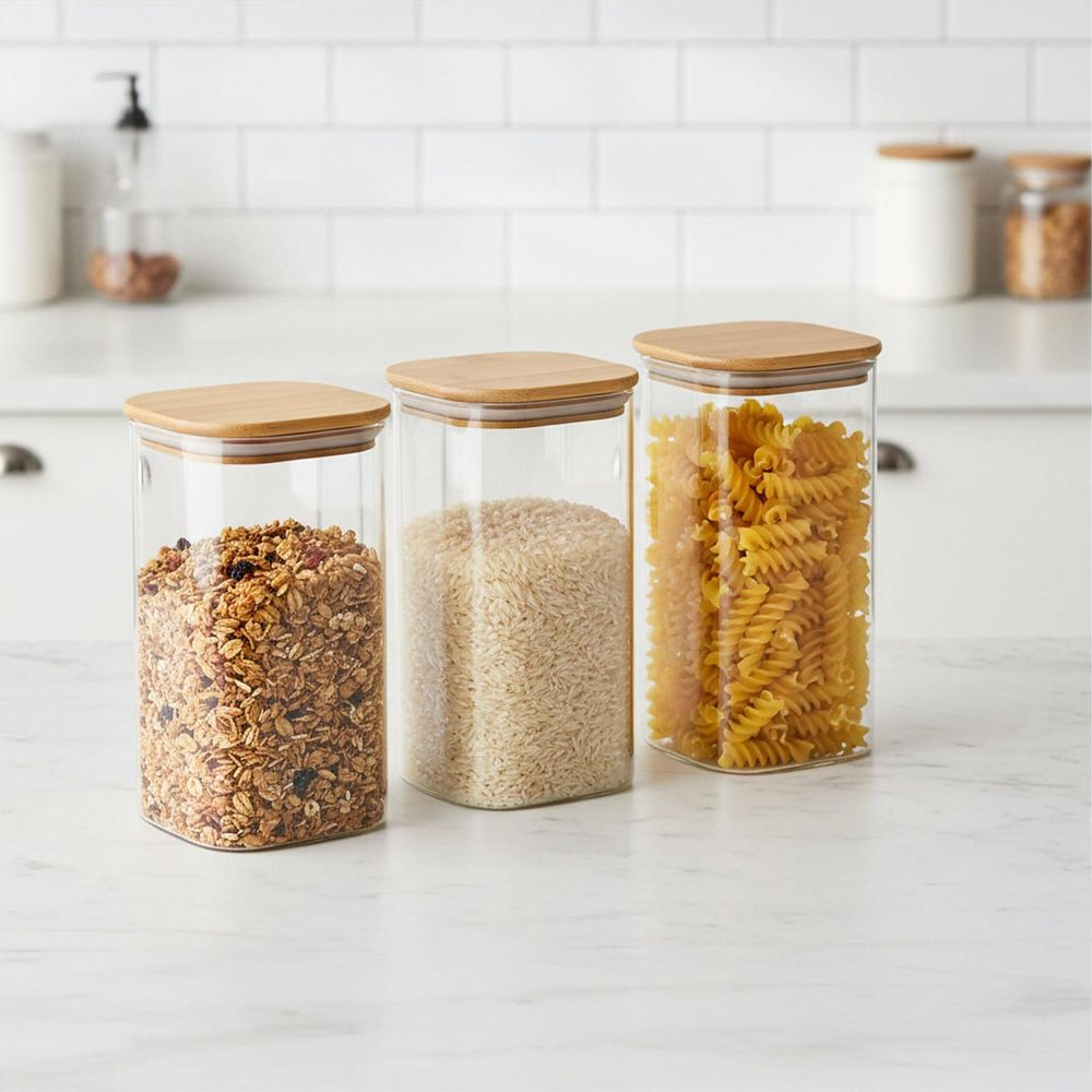 Drei Glasbehälter mit Holzdeckeln auf einer Küchenarbeitsplatte, gefüllt mit Müsli, Reis und Nudeln.