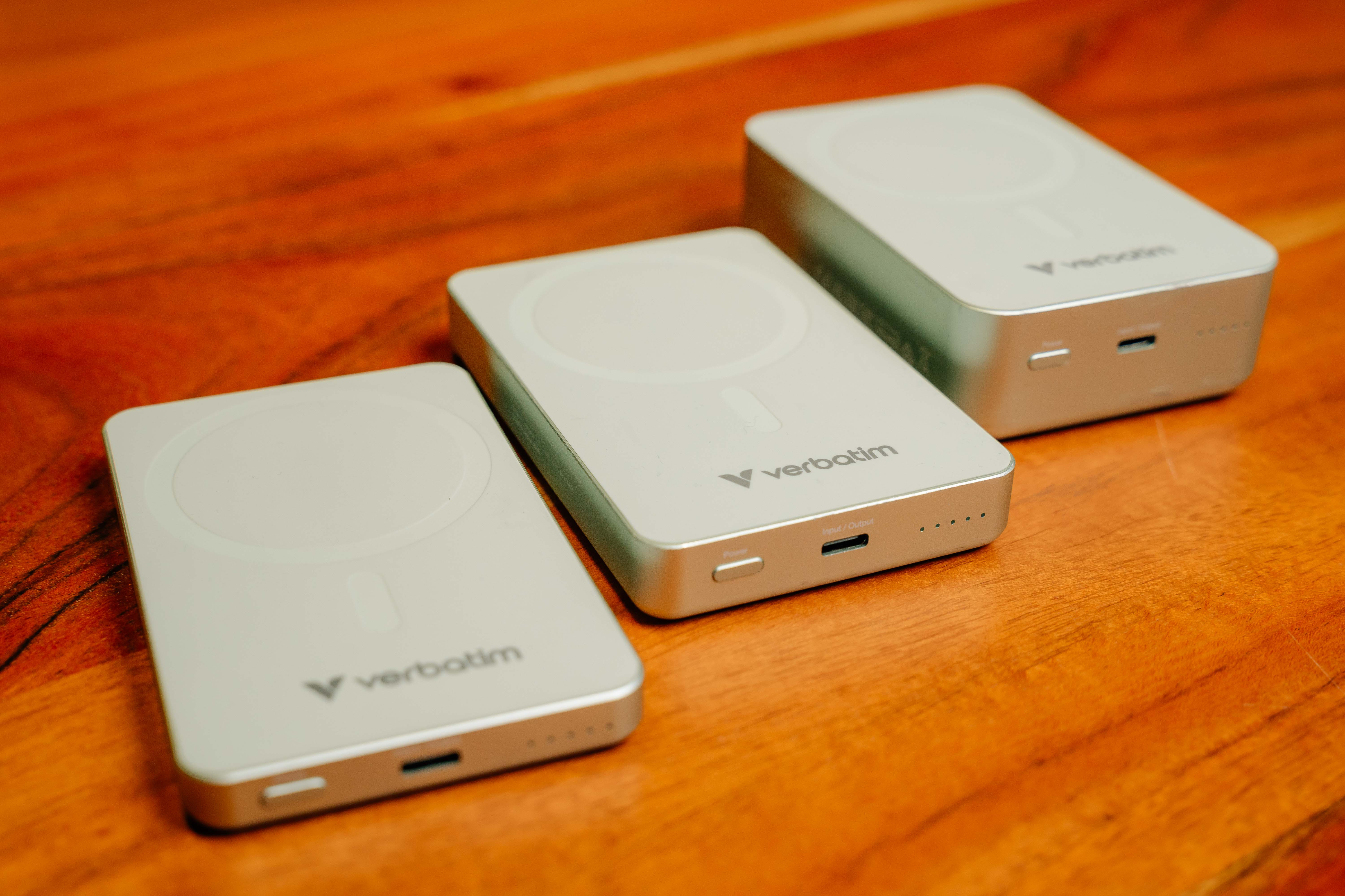 Drei silberne Verbatim Powerbanks diagonal auf einer Holzoberfläche angeordnet.