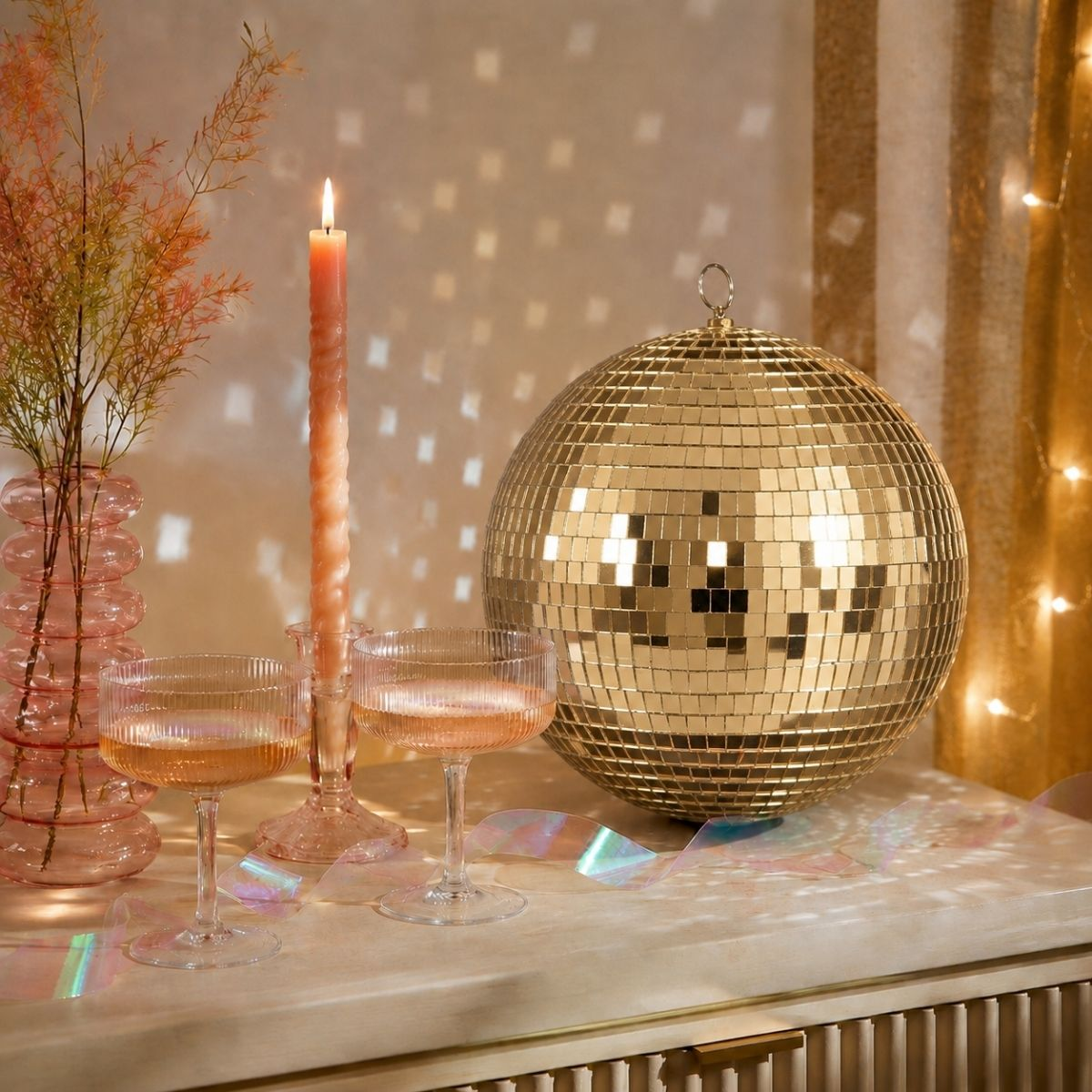 A gold disco ball, two champagne glasses, a pink candle, and a vase with branches on a table.
