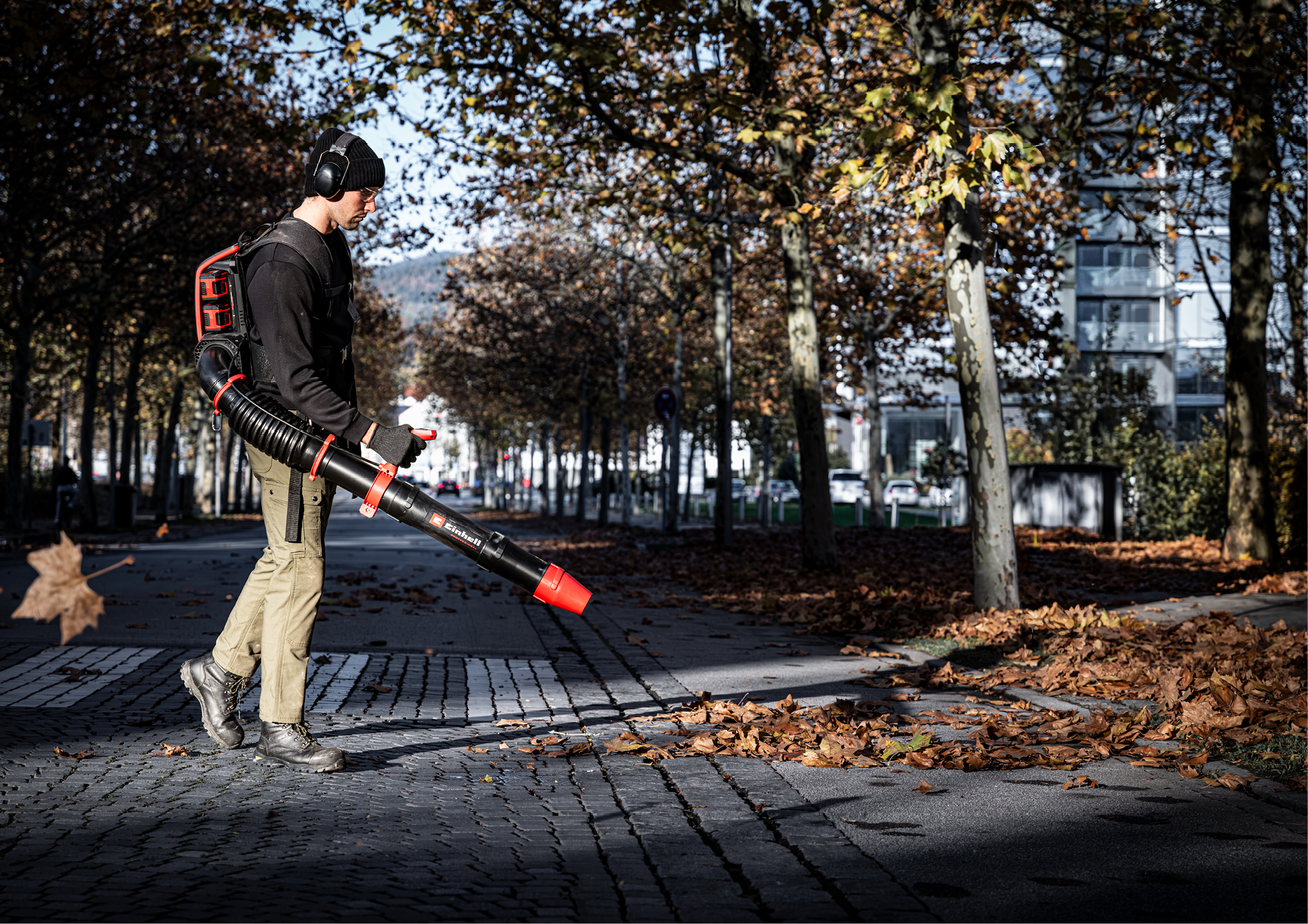 Ein Arbeiter nutzt einen Laubbläser auf einer herbstlichen Straße mit vielen abgefallenen Blättern.