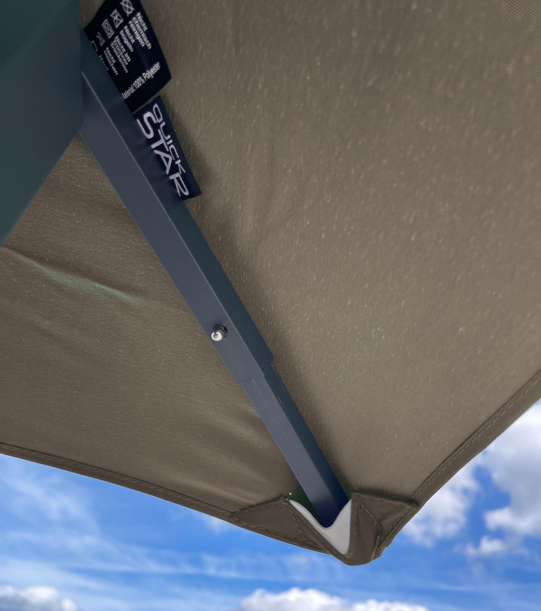 Unterseite eines braunen Sonnenschirms mit schwarzem Rahmen, Etikett und Metallstift gegen einen blauen Himmel.