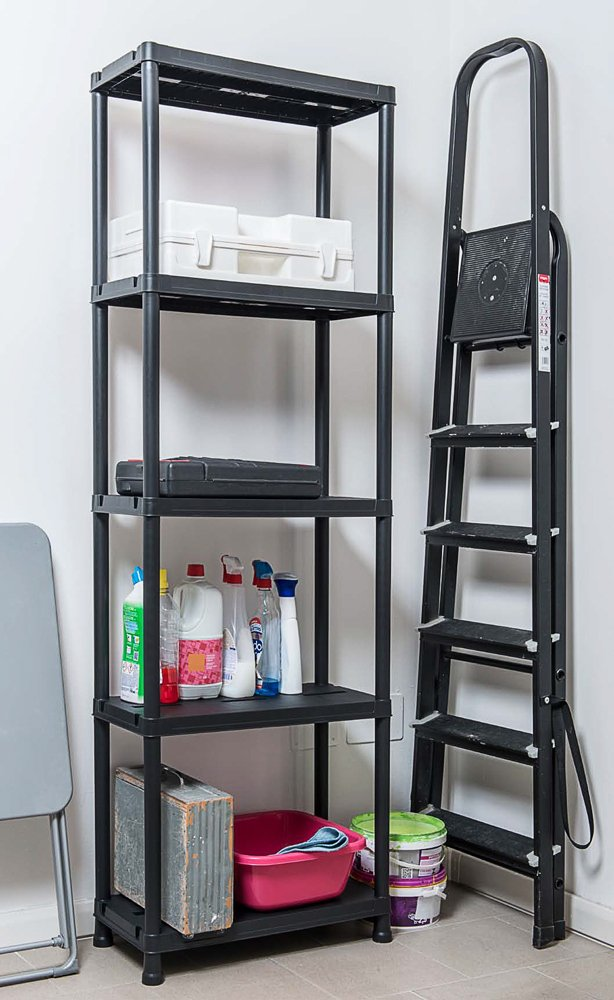 Ein schwarzes Kunststoffregal mit Reinigungsmitteln und Boxen steht neben einer schwarzen Leiter vor einer weißen Wand.