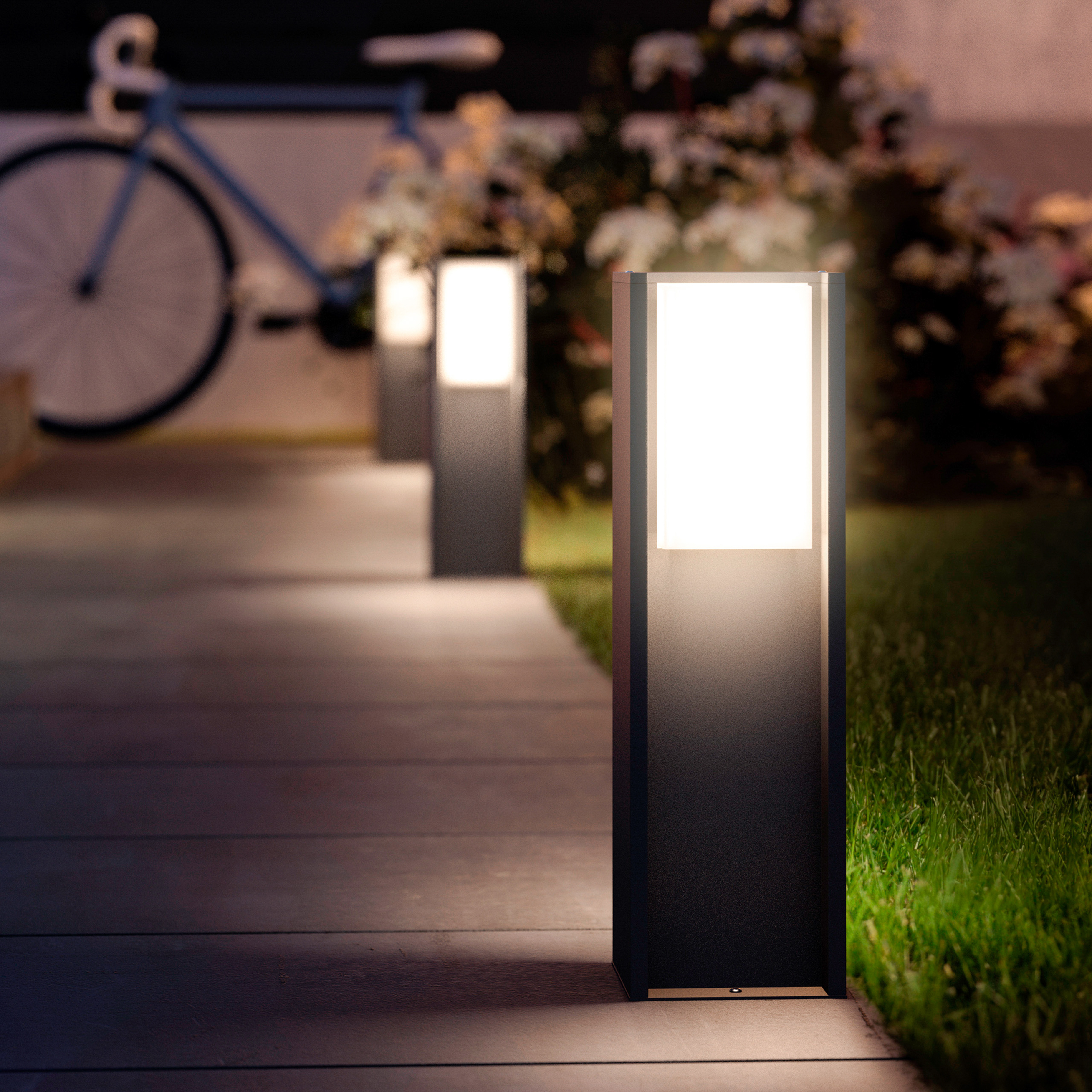 Luci da giardino illuminate illuminano un sentiero vicino al prato di notte, con una bicicletta blu sfocata.