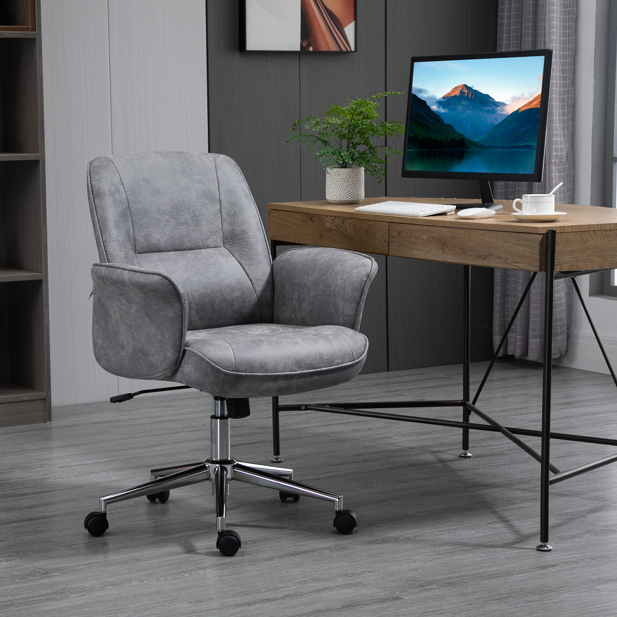 Una sedia da ufficio imbottita grigia con ruote si trova davanti a una scrivania in legno con monitor, pianta e tazza.
