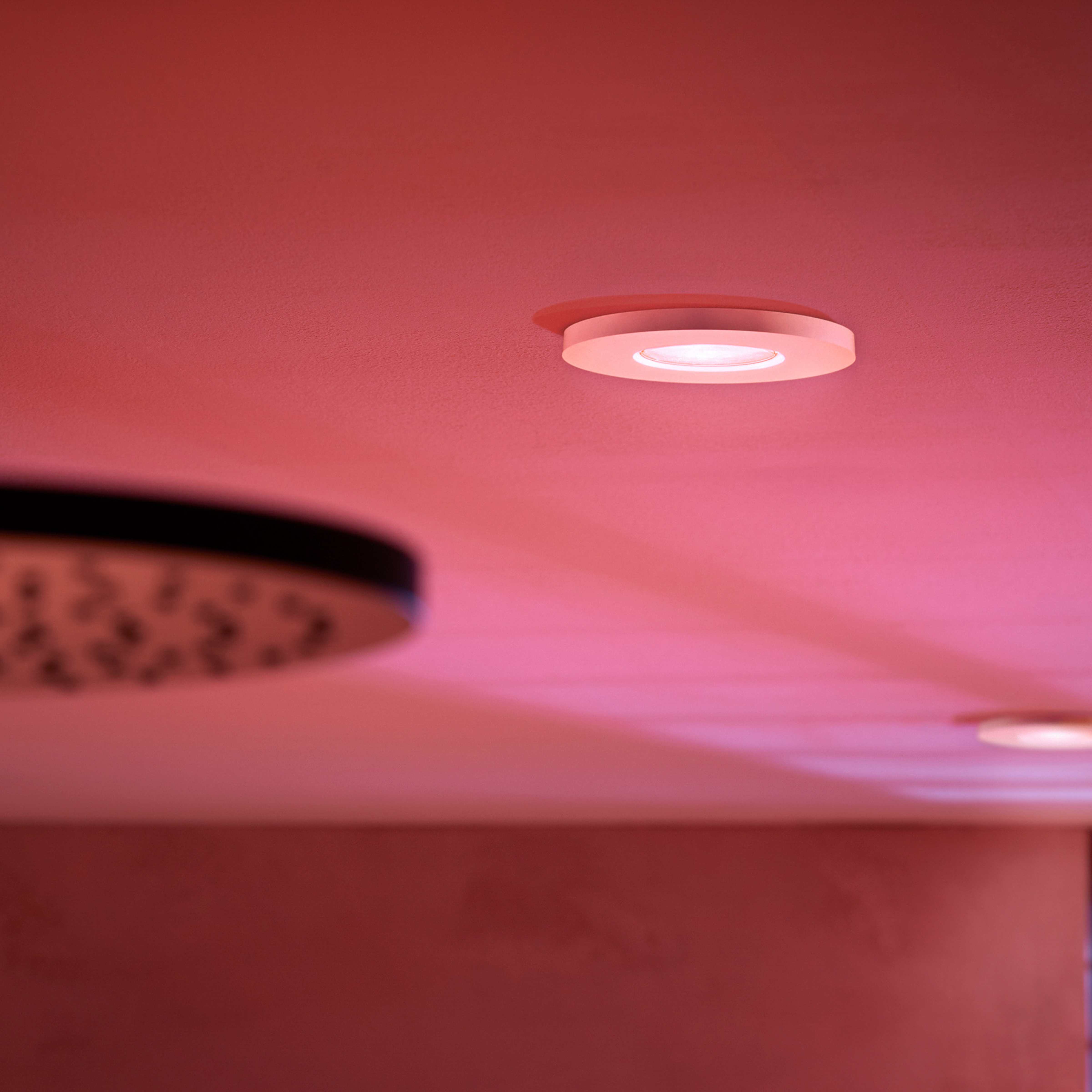 Una plafoniera a incasso illumina una stanza con una calda tonalità rosa, con un soffione sfocato in primo piano.