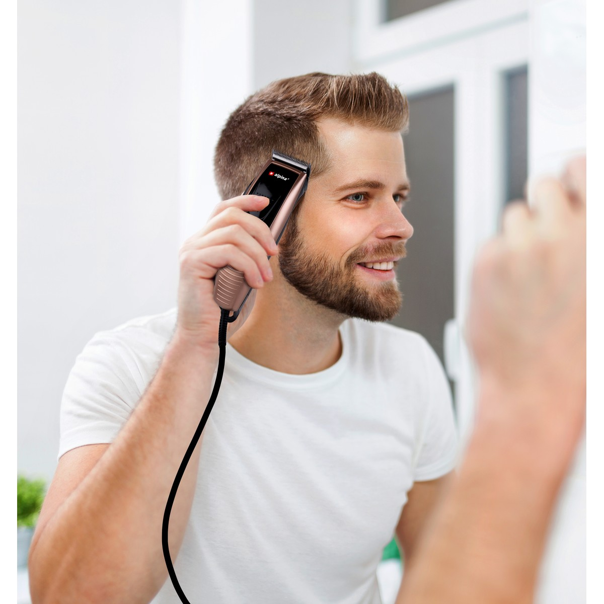 A man wearing a white t-shirt trims his hair in front of a mirror with a corded clipper.