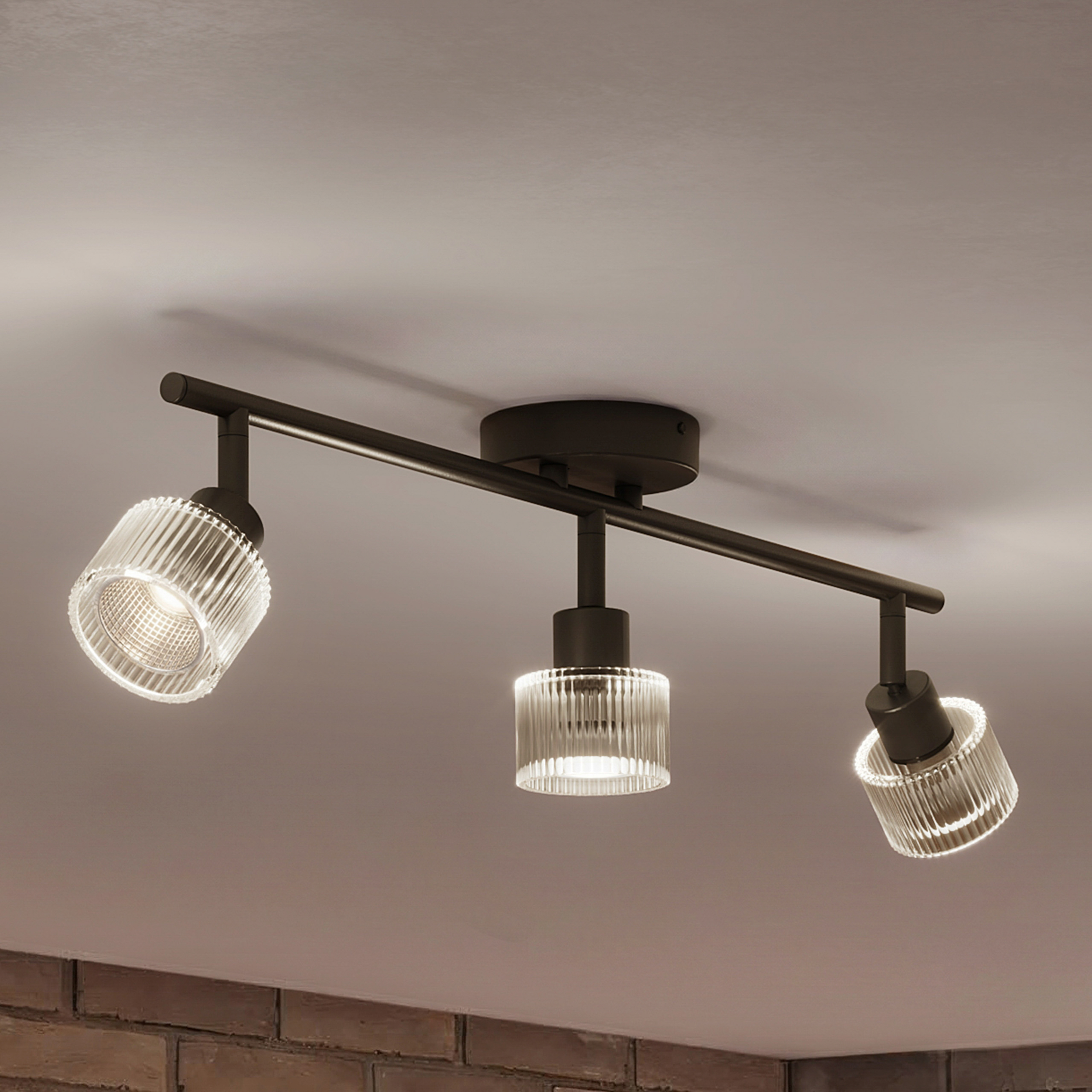 Eine schwarze Deckenleuchte mit drei gerippten Glasschirmen strahlt helles Licht gegen eine helle Decke.