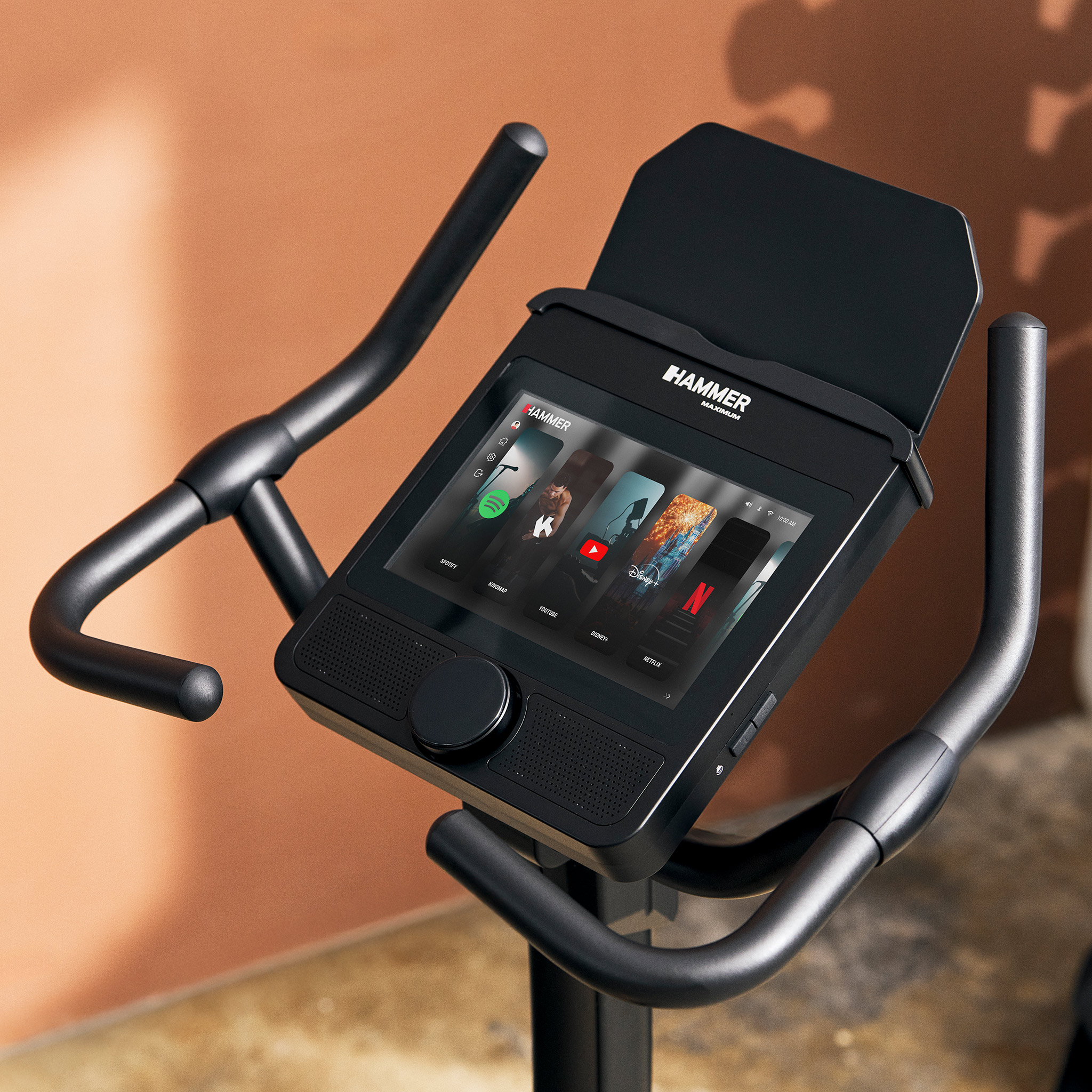 Nahaufnahme eines Heimtrainers mit digitalem Touchscreen und App-Symbolen vor warmem Hintergrund.