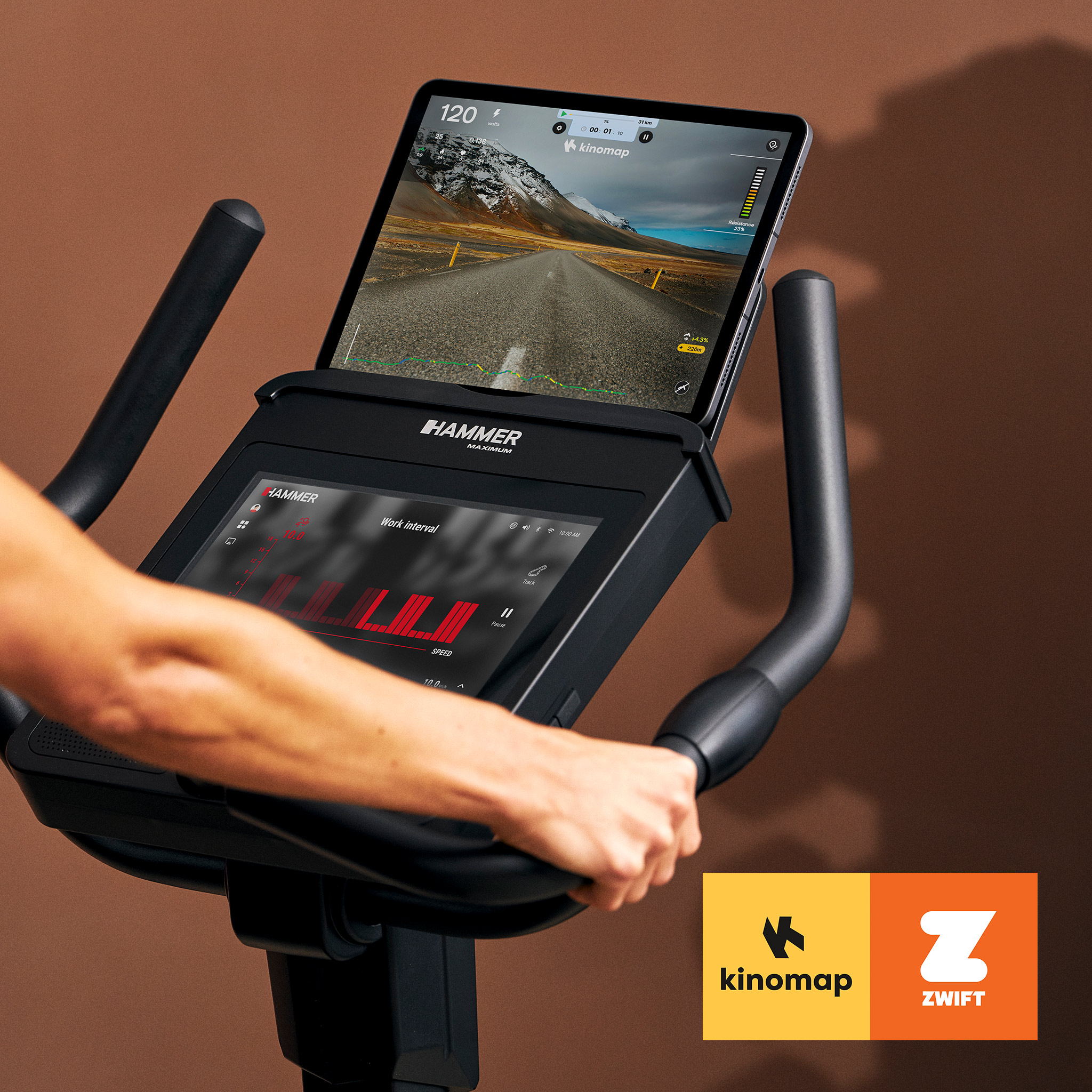 Person hält den Lenker eines Heimtrainers mit Tablet und Kinomap-App vor brauner Wand.