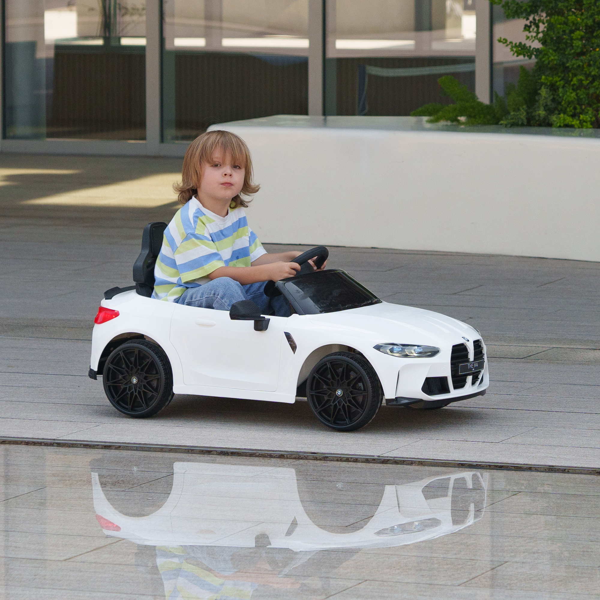 Un bambino biondo siede su un'auto giocattolo bianca su pavimento piastrellato con un riflesso sotto.