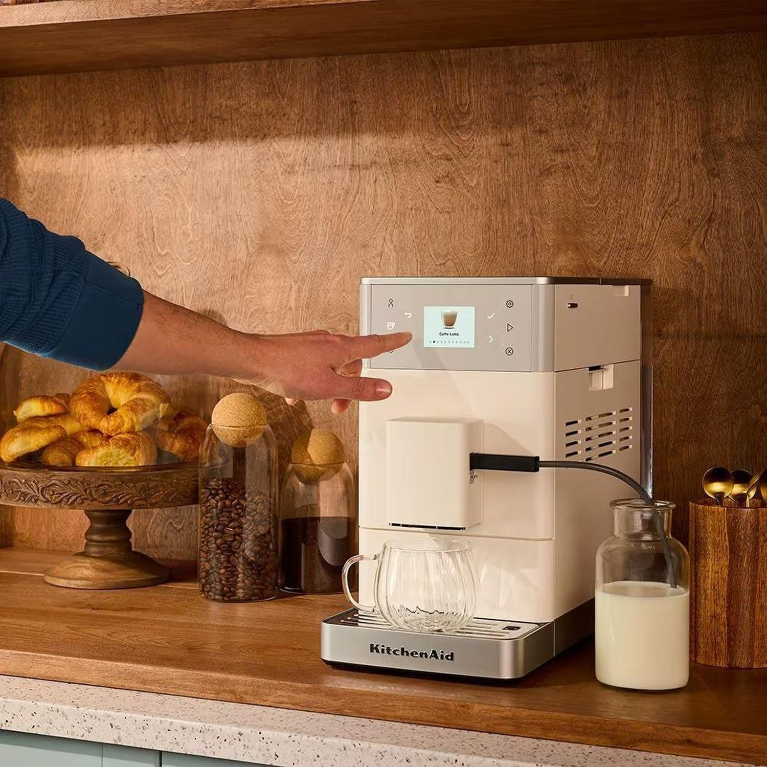 Ahşap tezgah üzerinde süt şişesi ve kruvasanların yanında beyaz KitchenAid kahve makinesini çalıştıran kişi.