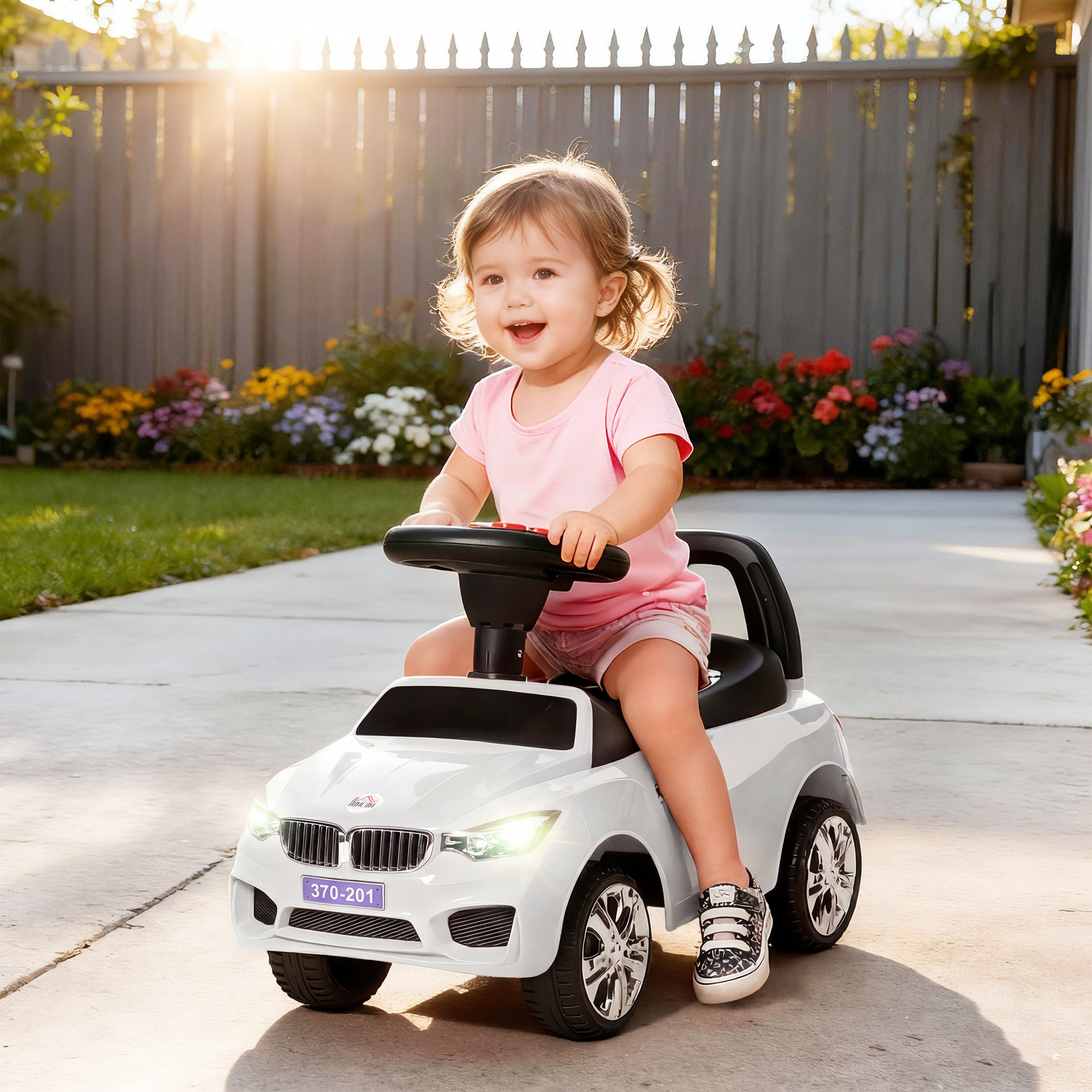 Bambino sorridente con maglietta rosa su un'auto giocattolo bianca in un giardino.