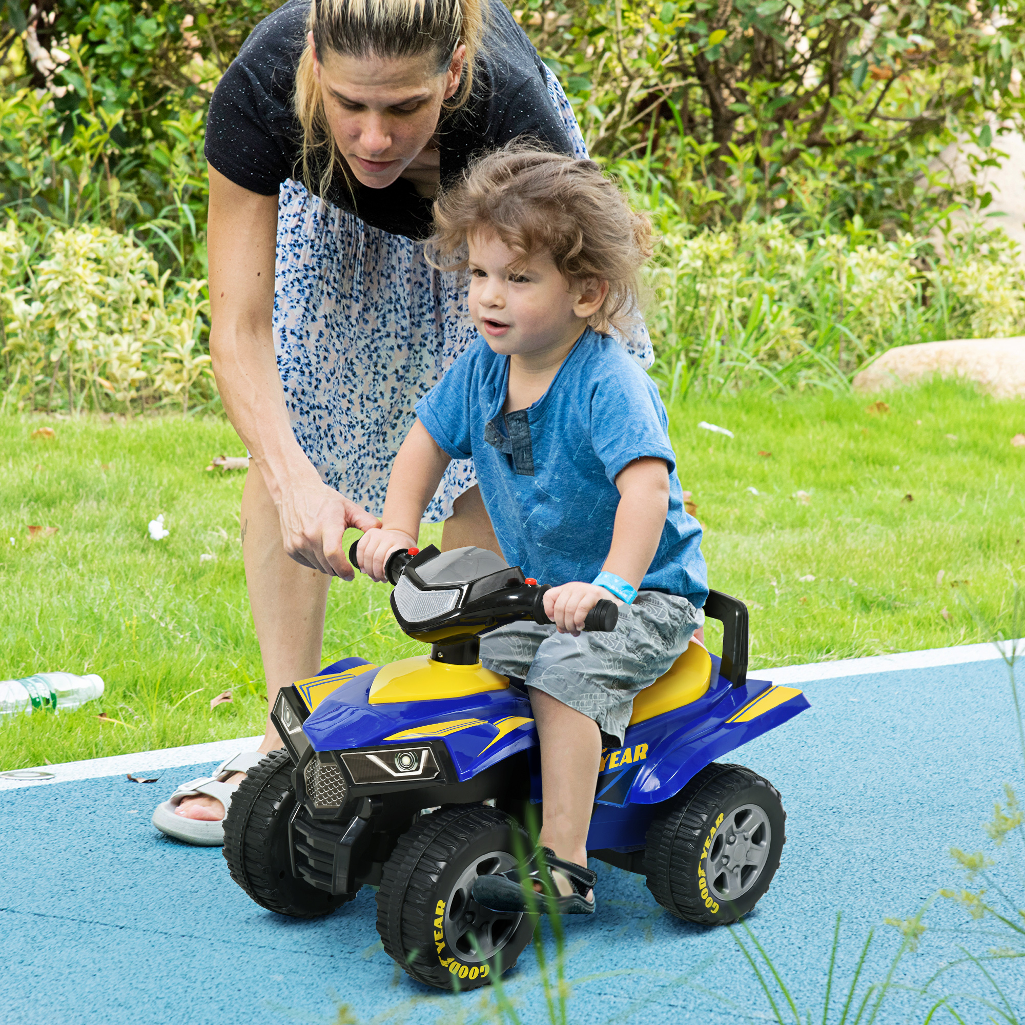 Un bambino guida un quad giocattolo blu all'aperto con una donna in piedi dietro di lui.