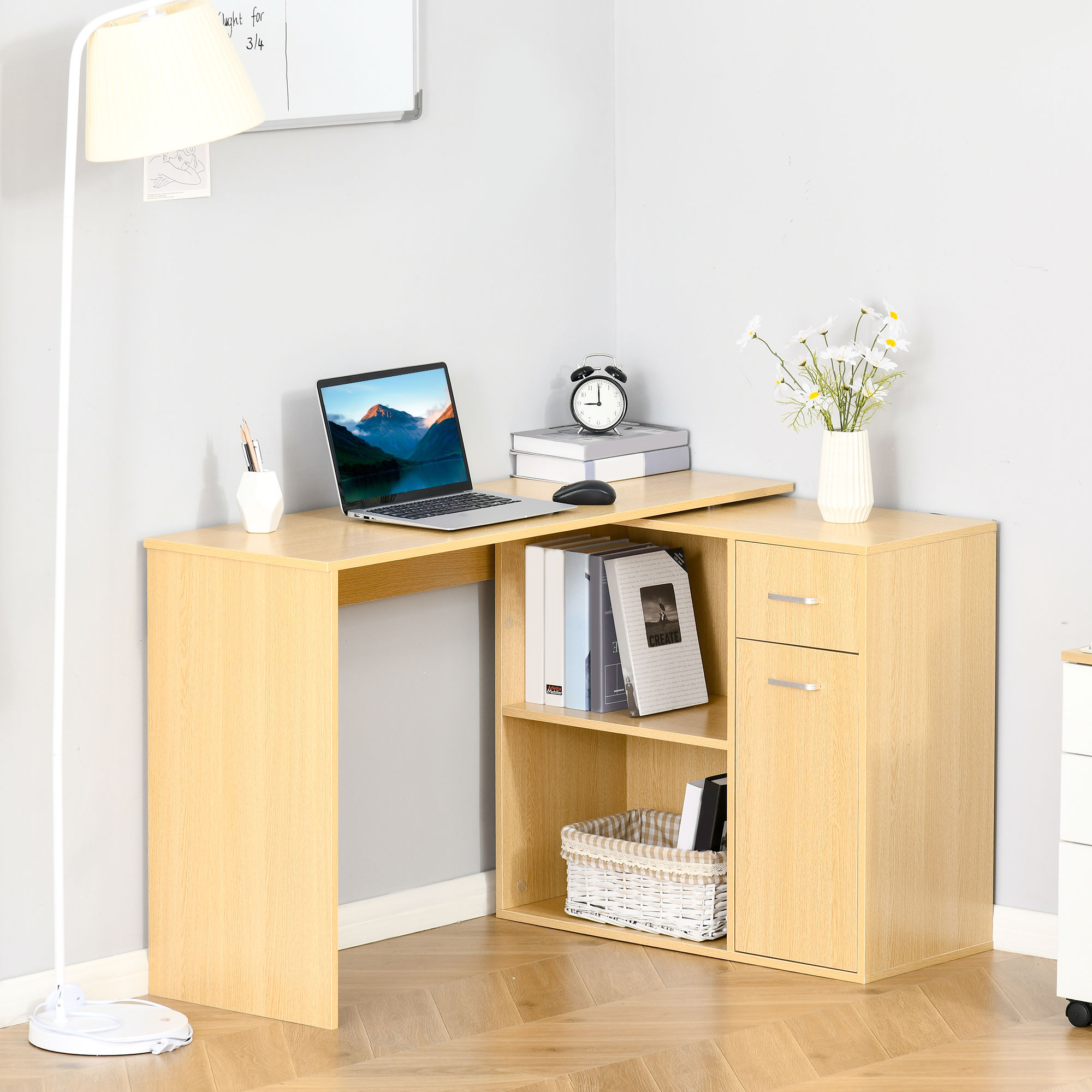 Scrivania in legno chiaro a L con laptop, libri, fiori e lampada da terra.