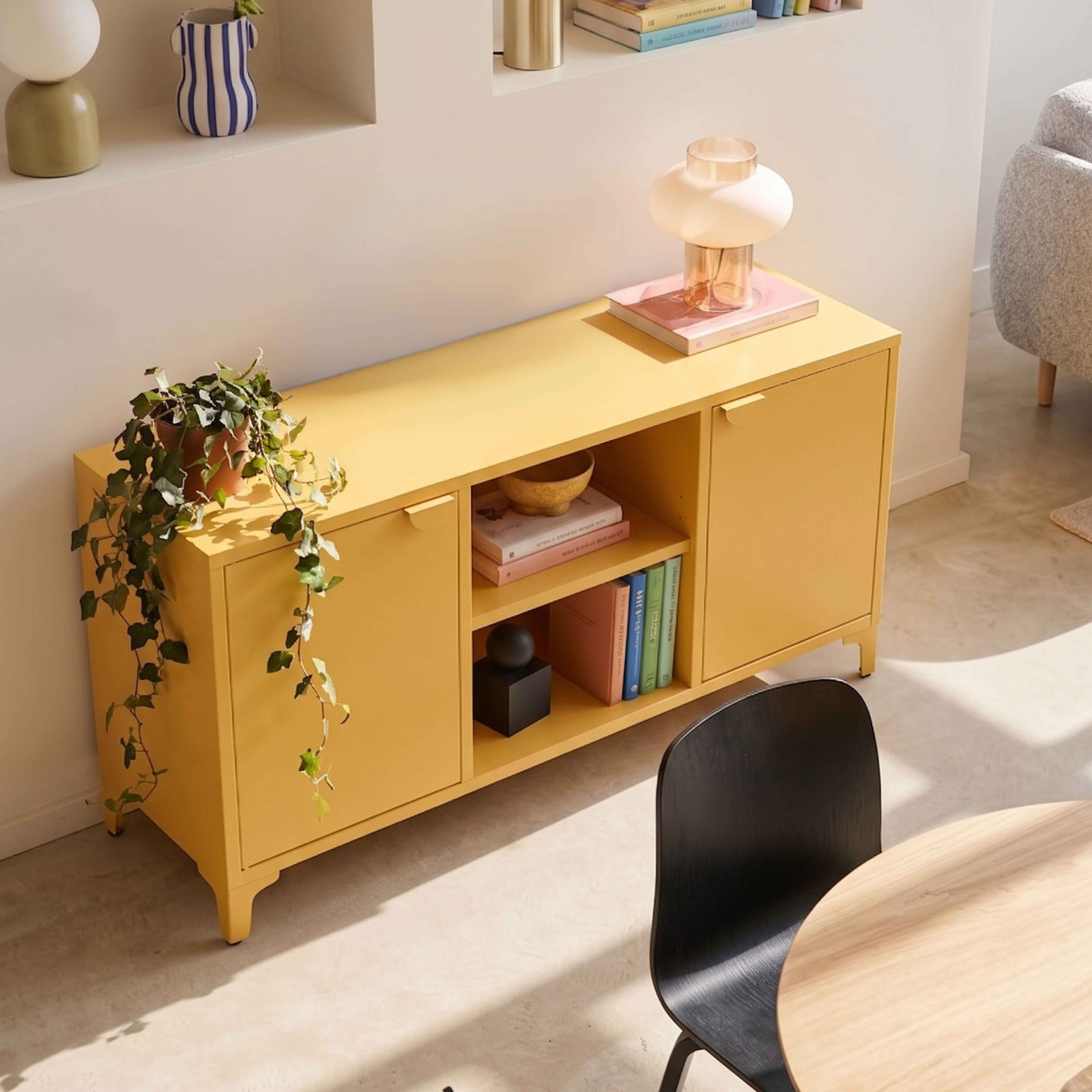 Ein gelbes Sideboard mit Pflanzen, Büchern und einer Lampe steht an einer Wand neben einem Tisch.