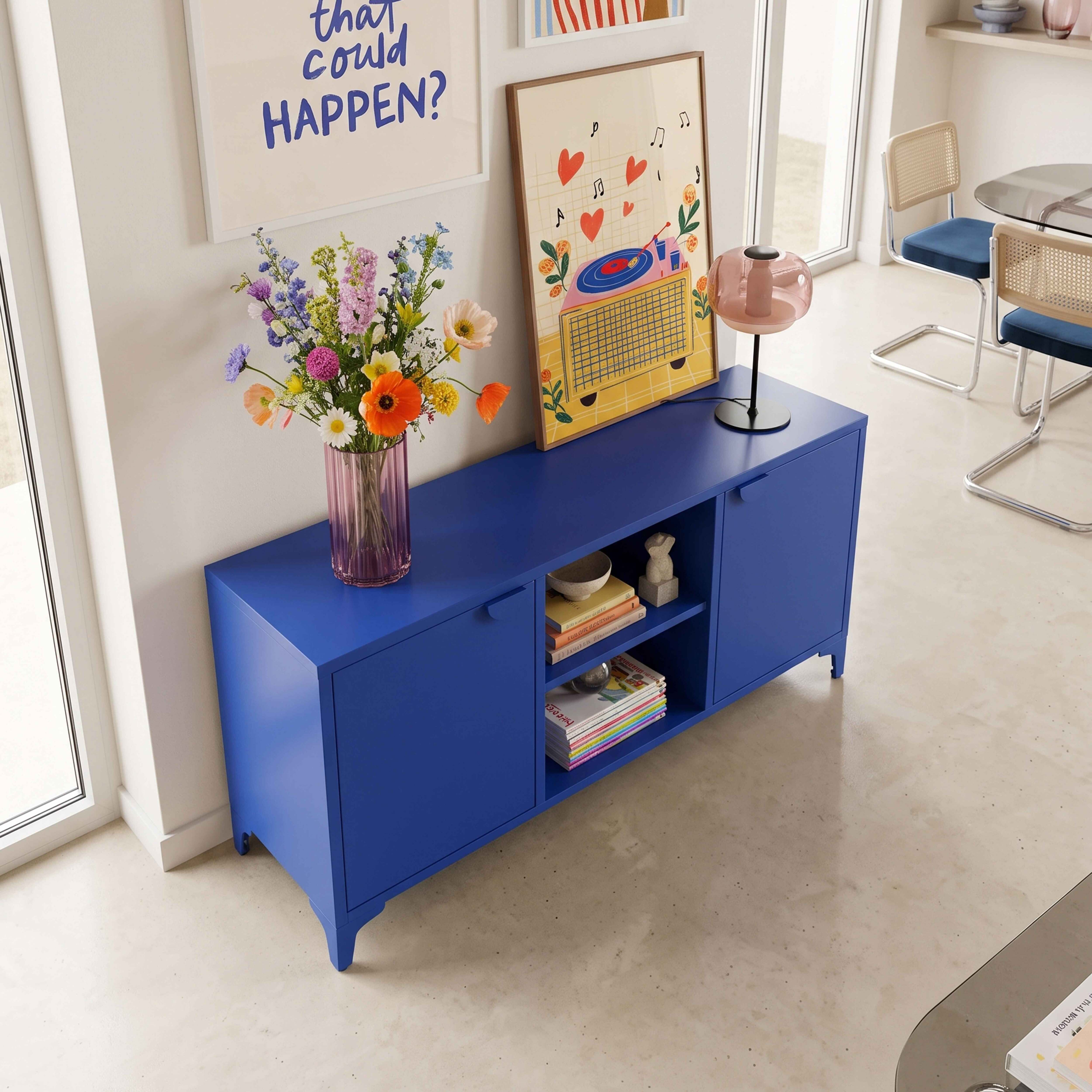 Ein leuchtend blauer Anrichteschrank trägt eine Vase mit bunten Blumen, Büchern und eine Lampe vor einer hellen Wand.