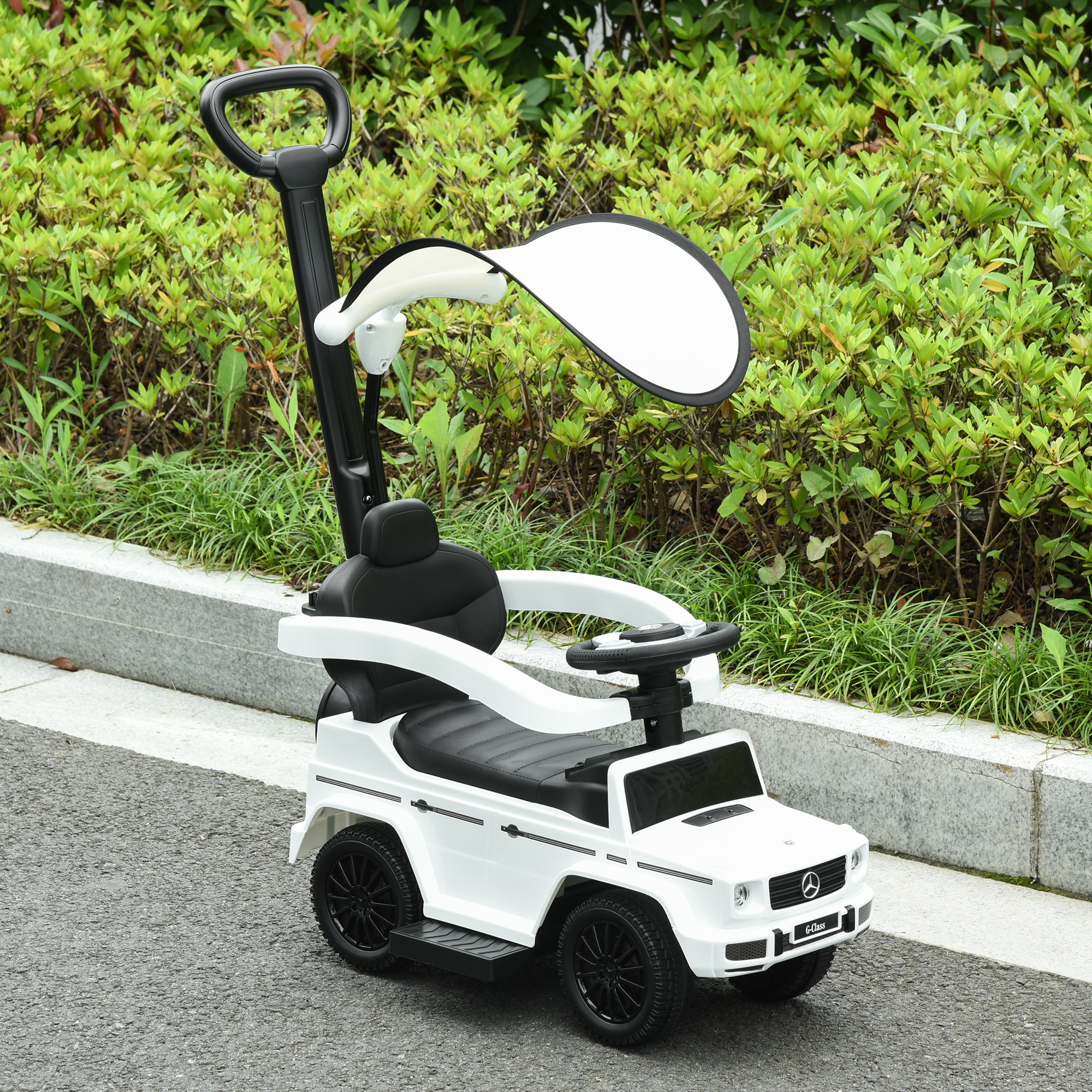 Un'auto giocattolo cavalcabile bianca con maniglia e tendalino parasole parcheggiata su un sentiero vicino a cespugli.
