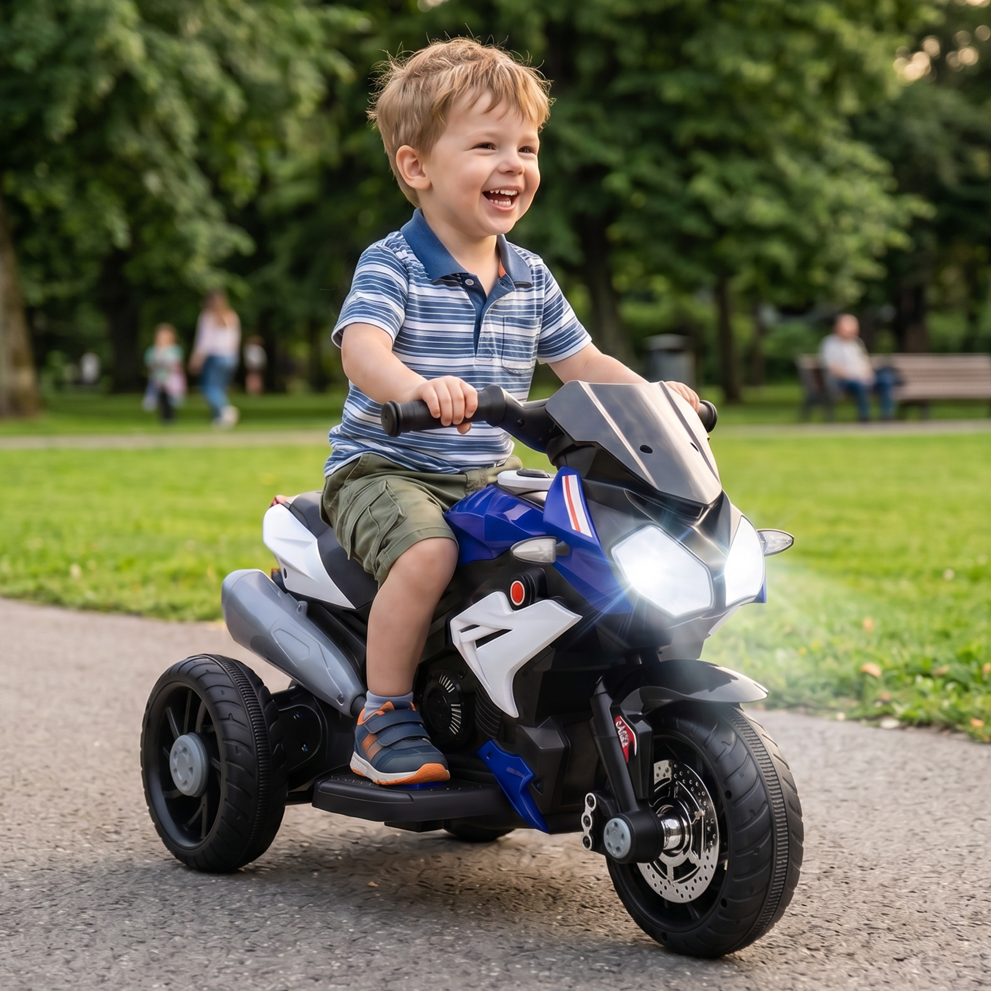 Un bambino sorridente guida una moto giocattolo blu e bianca su un sentiero asfaltato in un parco verde con alberi.
