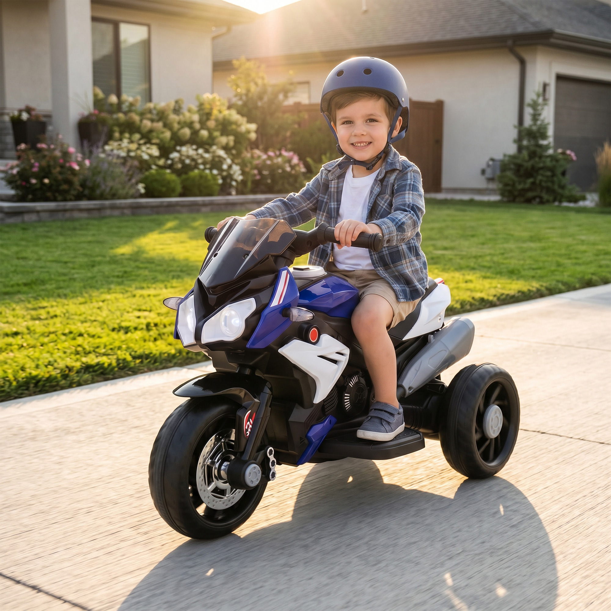 Un bambino sorridente con casco blu guida una moto giocattolo a tre ruote su un vialetto soleggiato davanti a una casa.
