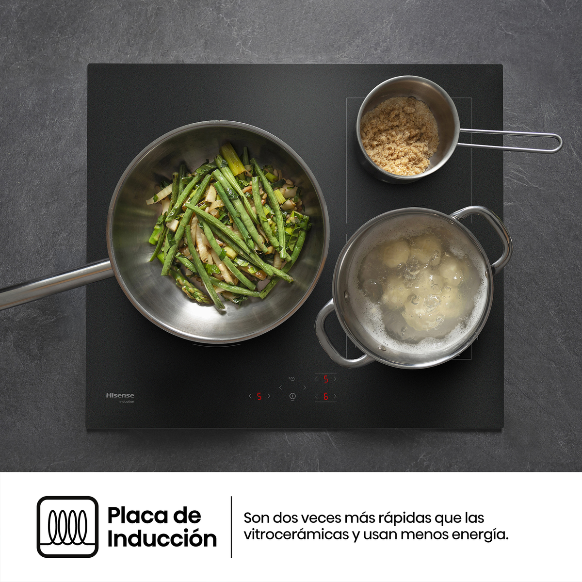 Placa de inducción negra con ollas y sartén con verduras, fotografiada desde arriba sobre una encimera oscura.