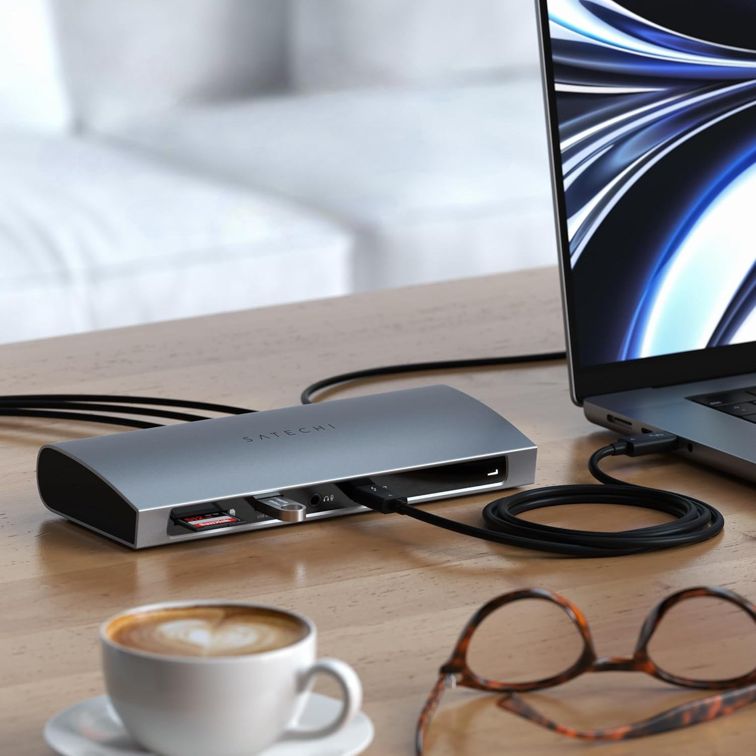 Eine graue Satechi-Dockingstation auf einem Holztisch neben einem Laptop, einer Kaffeetasse und einer Brille.