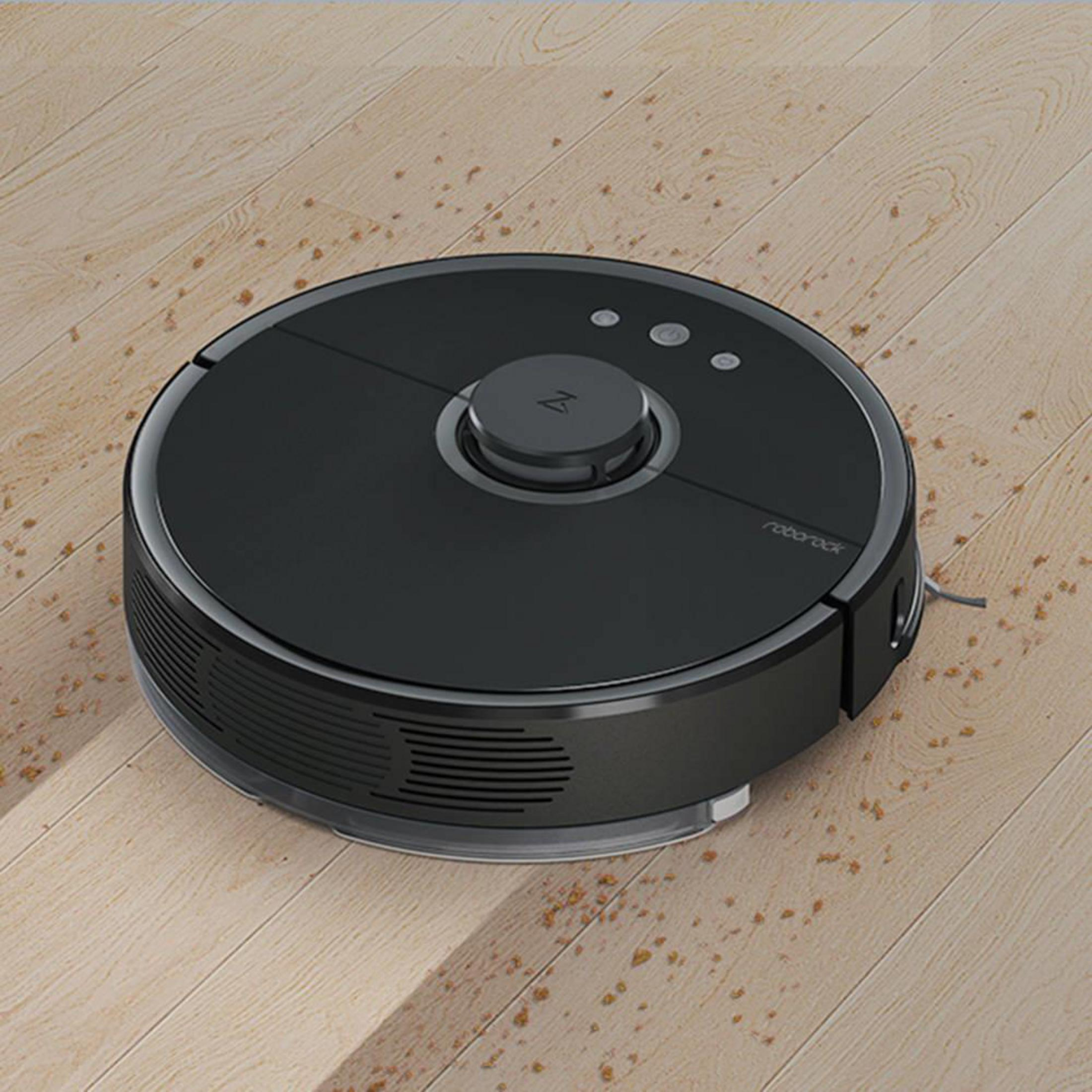 Ein schwarzer Staubsaugerroboter auf einem Holzboden mit verstreutem Schmutz.