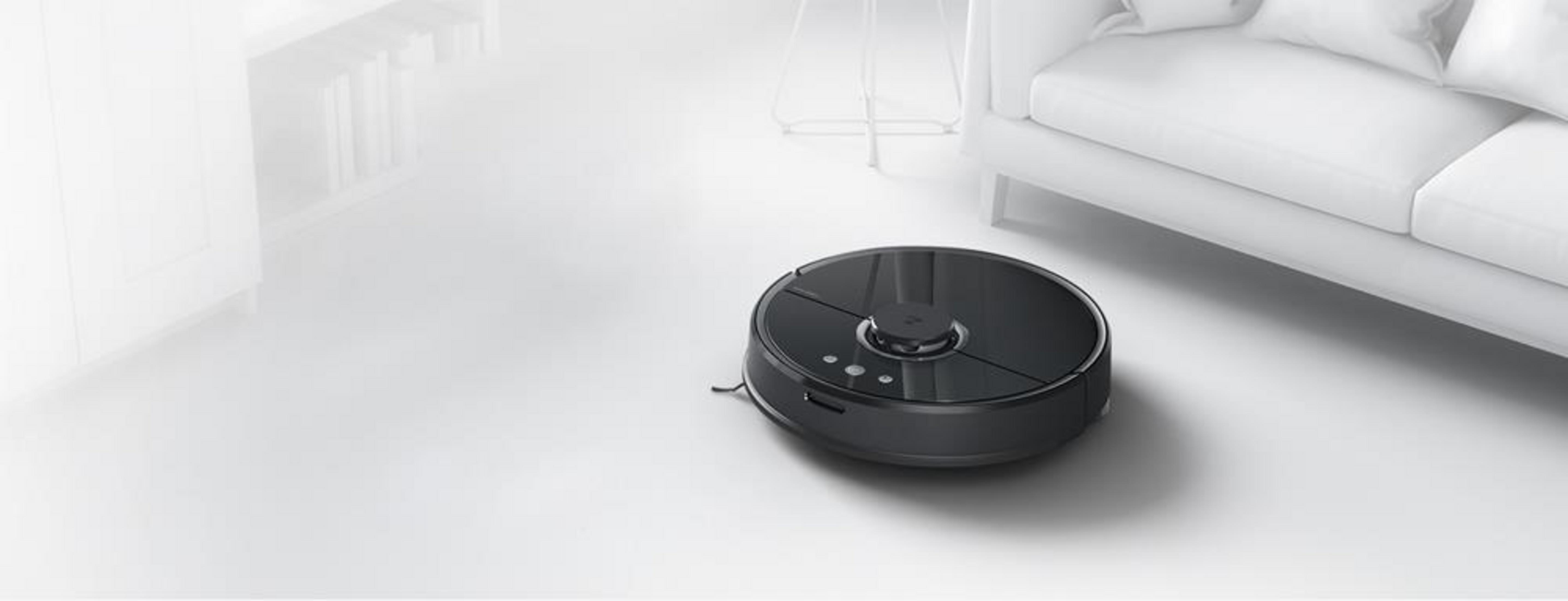 Ein schwarzer Saugroboter steht auf einem weißen Boden in einem hellen, modernen Wohnzimmer vor einem Sofa.