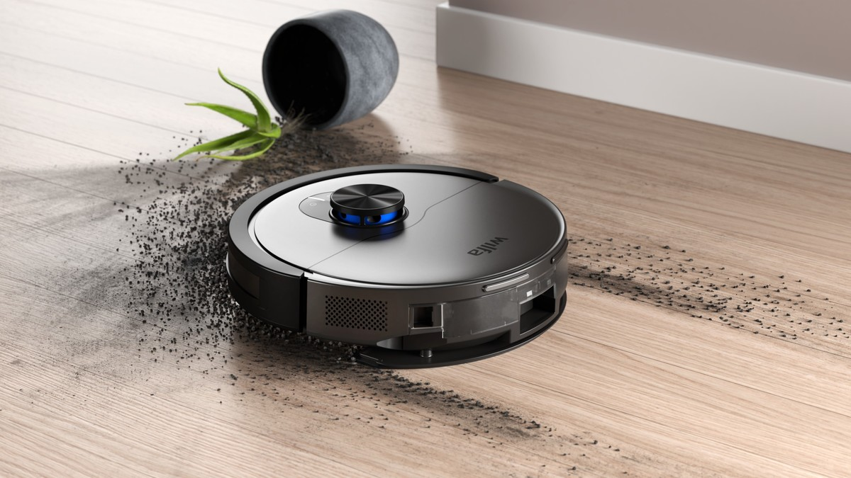 Un robot aspirapolvere nero pulisce la terra di un vaso rovesciato sul pavimento in legno.