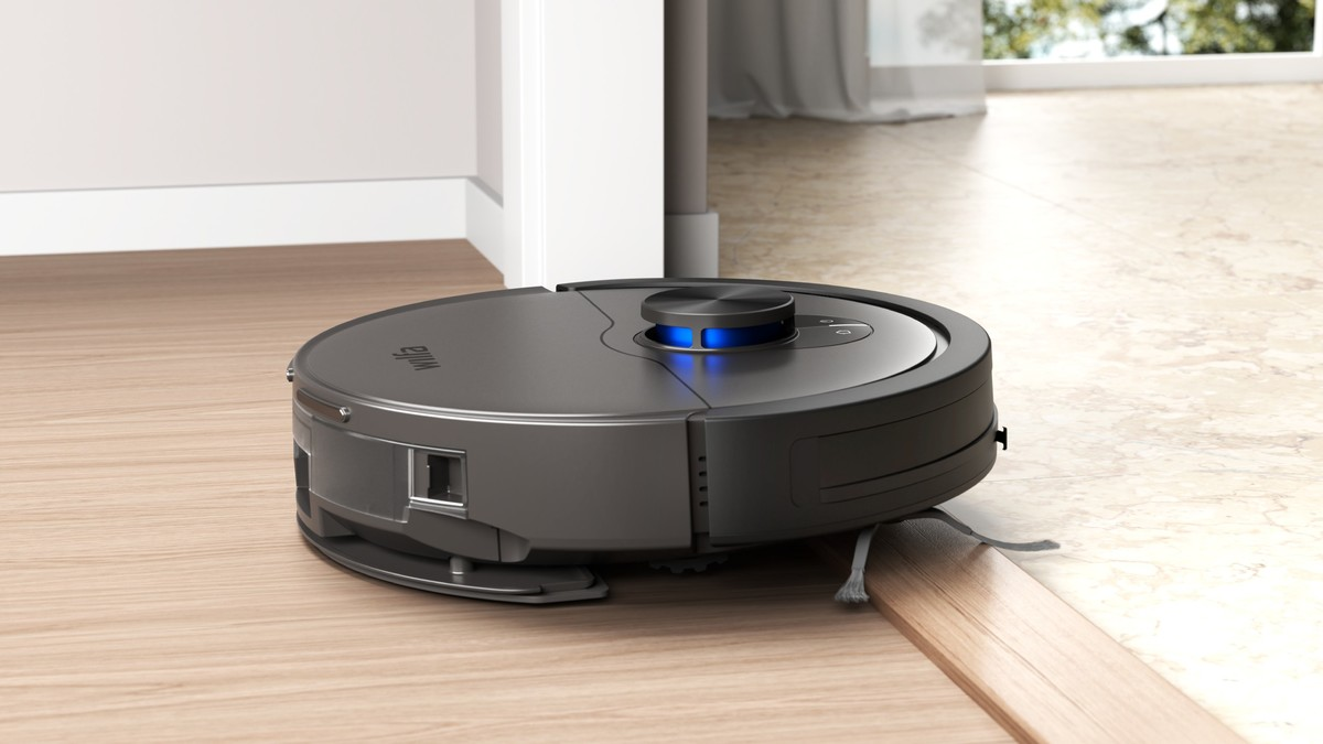 Un robot aspirapolvere grigio rotola su pavimento in legno oltrepassando una soglia.