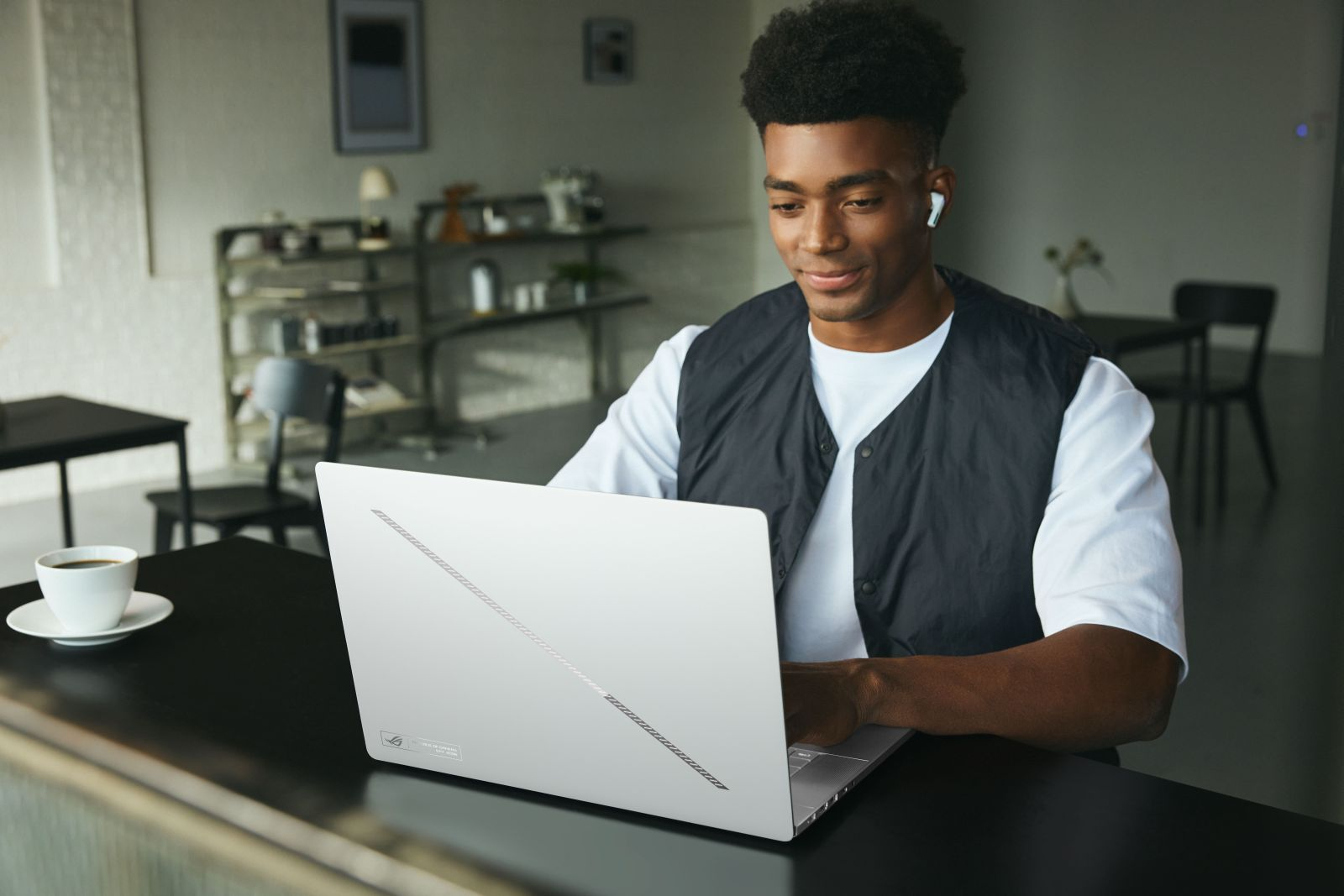 Ein junger Mann mit Weste und Ohrhörern sitzt in einem Café an einem Tisch und arbeitet an einem weißen Laptop.