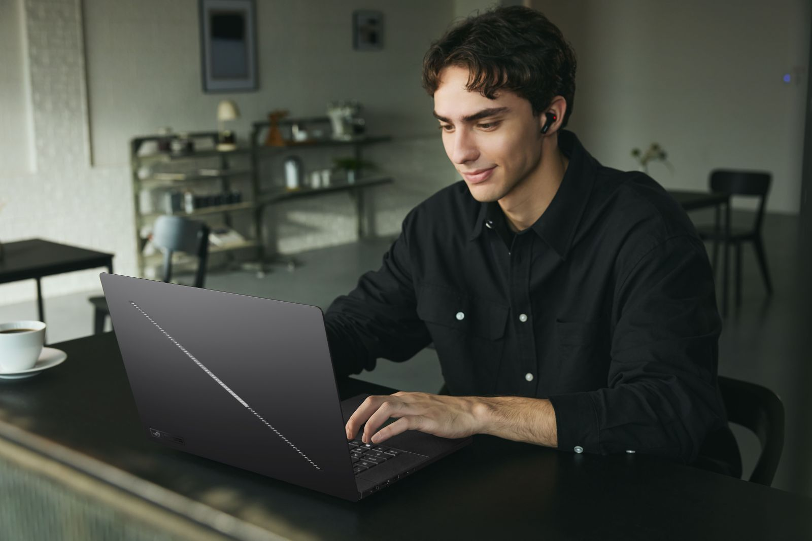 Ein junger Mann im schwarzen Hemd sitzt in einem Café und tippt lächelnd auf einem schwarzen Laptop.