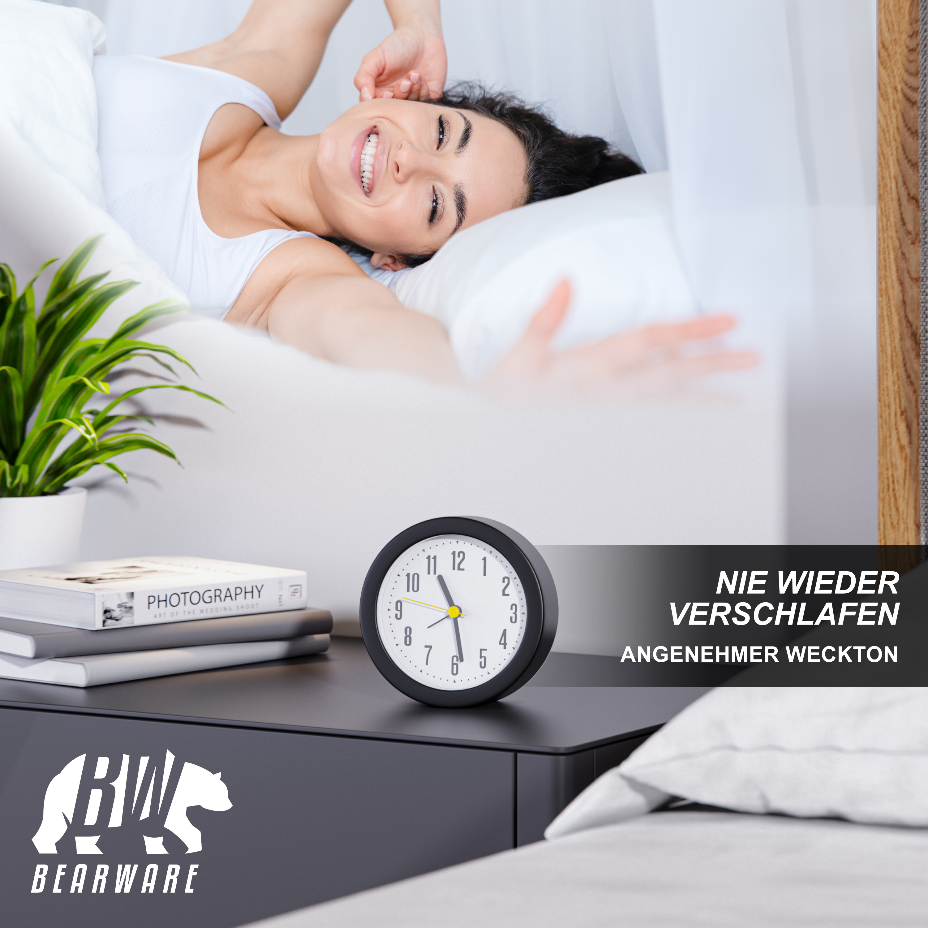 Ein Wecker und Bücher auf einem Nachttisch, während eine glückliche Frau im Bett aufwacht.