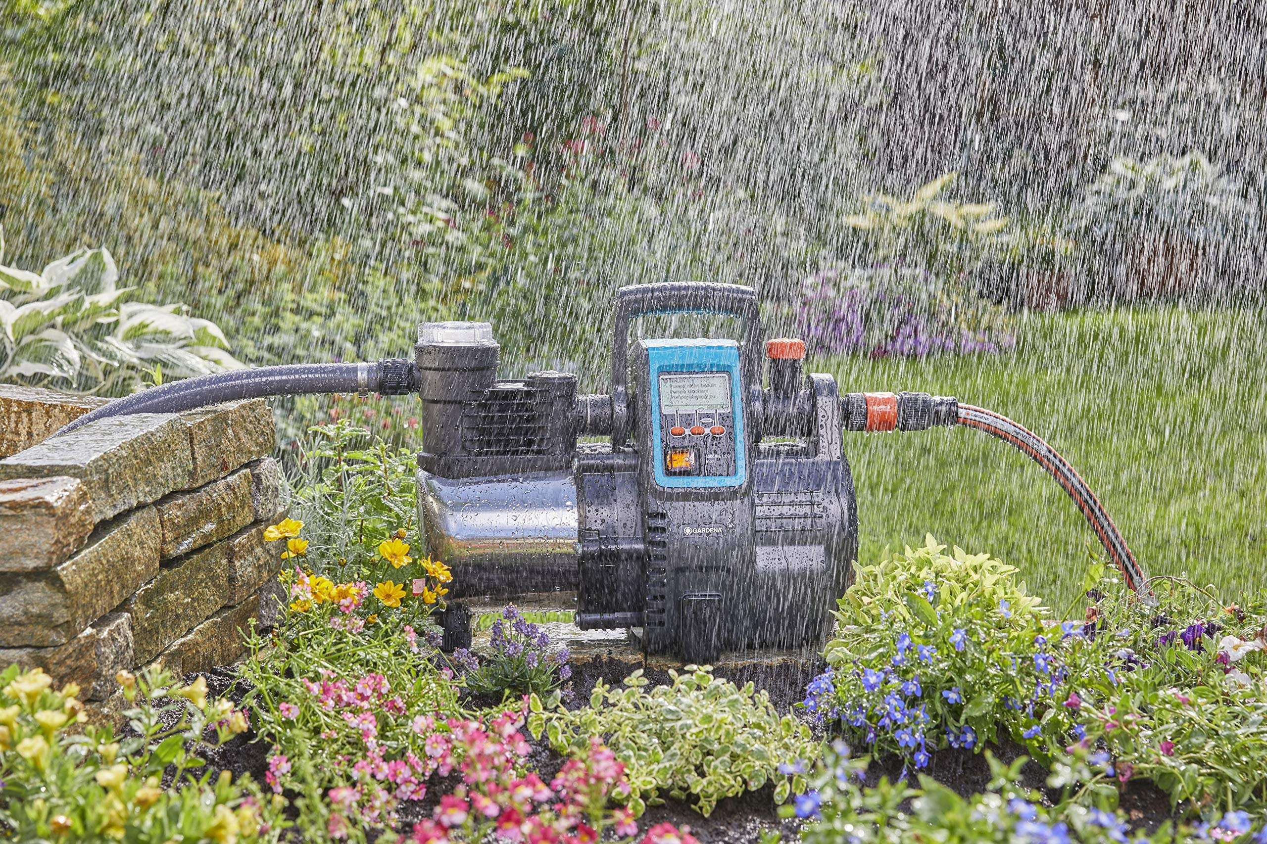 Eine Gartenpumpe steht in einem Garten, während es im Vordergrund stark regnet, umgeben von bunten Blumen.