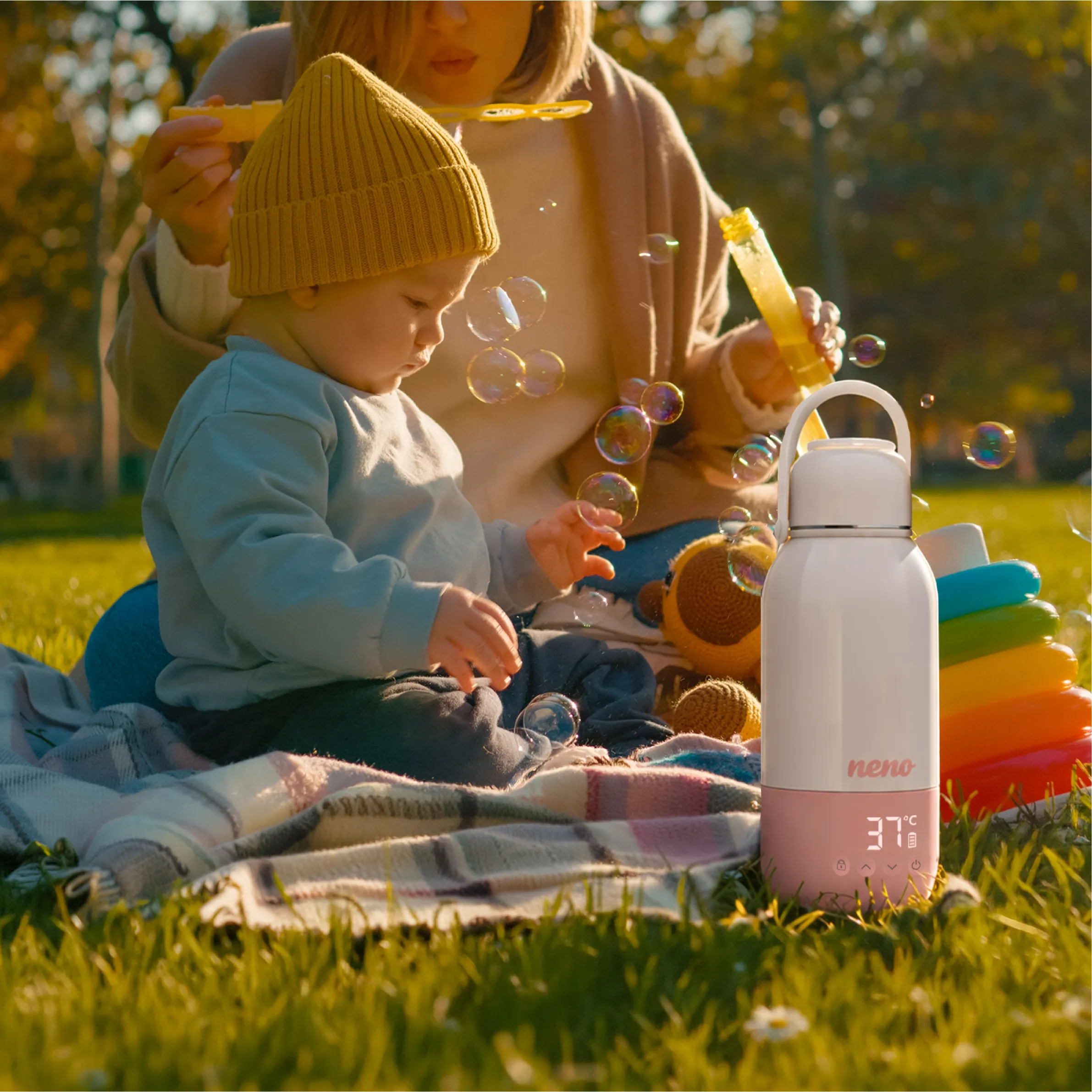 Ein Kleinkind mit gelber Mütze und eine Frau blasen Seifenblasen in einem Park, vorne eine weiße und rosa Neno-Flasche.