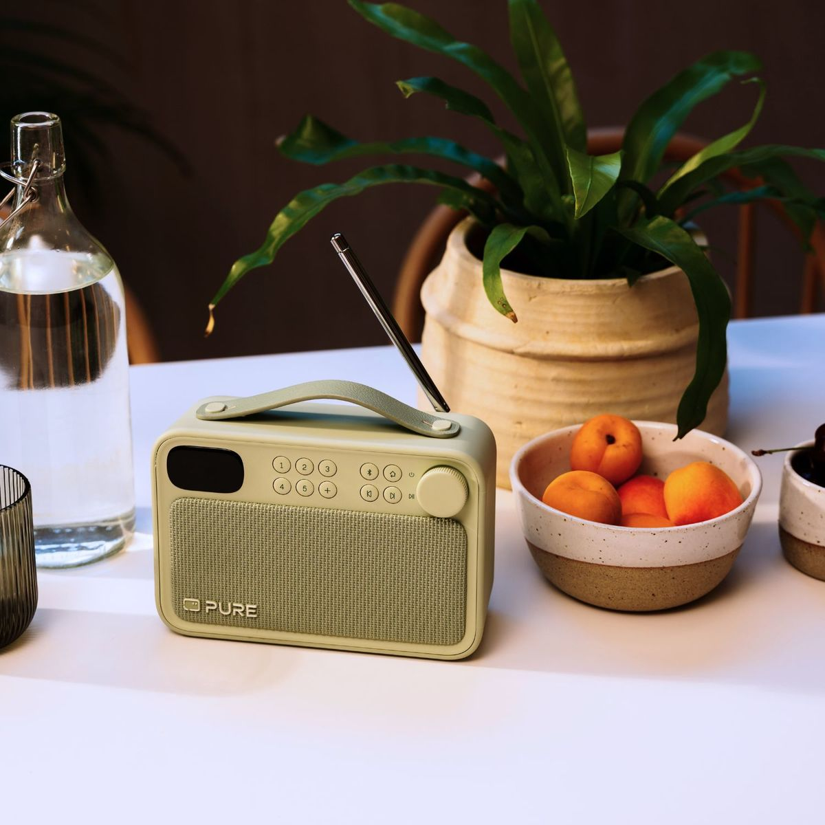 Una radio Pure verde claro en una mesa junto a una botella de agua, un bol de albaricoques y una planta en maceta.