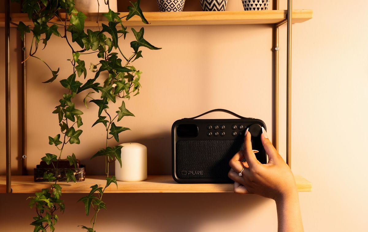 Una mano gira el dial de una radio Pure negra en un estante de madera junto a una vela blanca y hiedra.