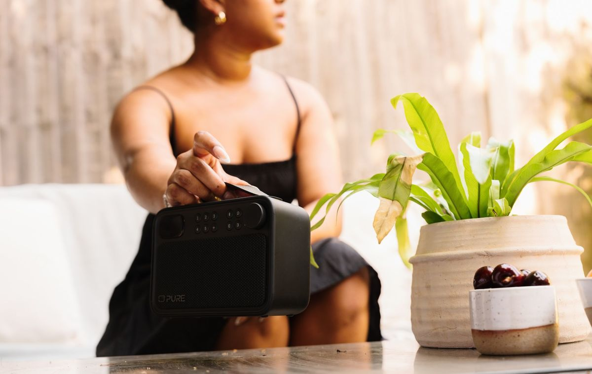 Una mujer sostiene una radio negra cerca de una planta en maceta con hojas verdes.
